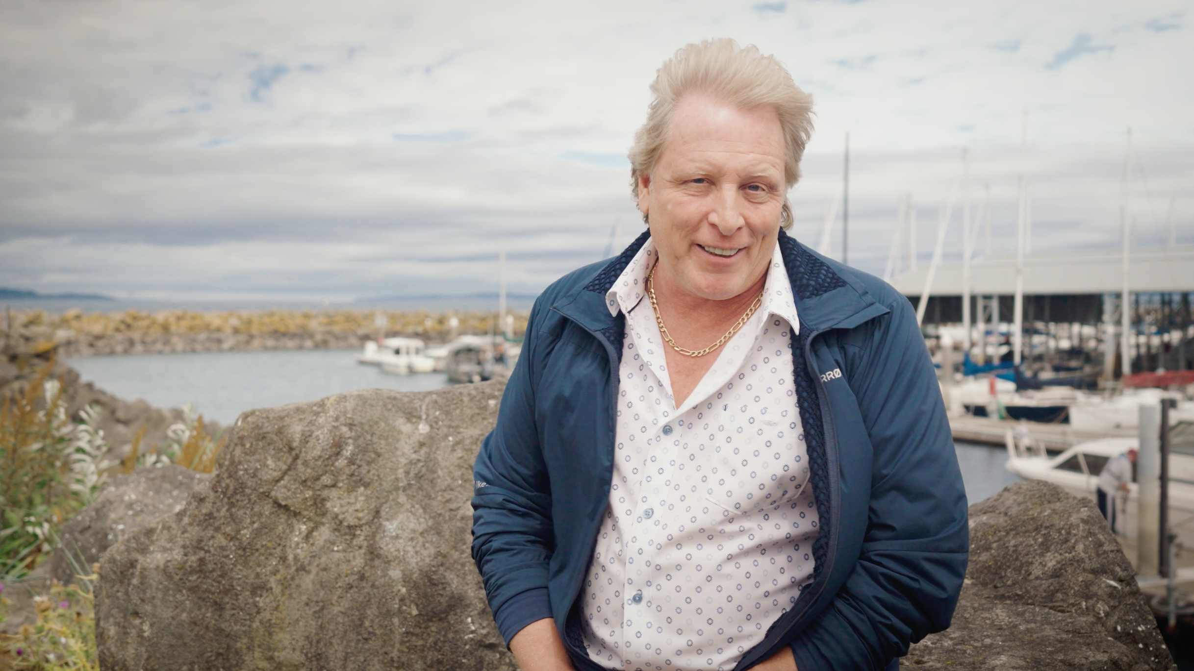 A man with light-colored hair smiling outdoors by a marina with boats, rocks, and cloudy sky in the background.