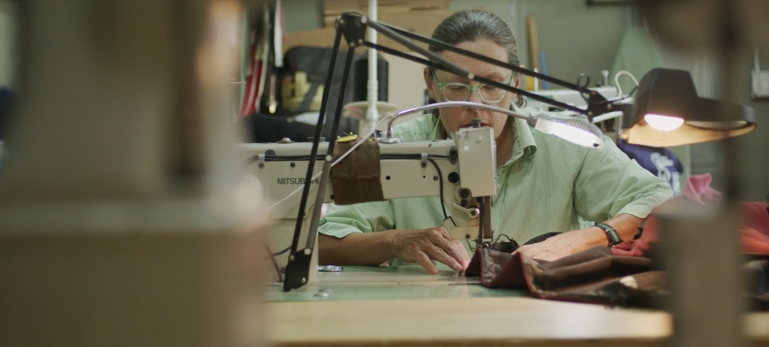 A person working on a sewing machine, wearing glasses and a green shirt, in a workshop.