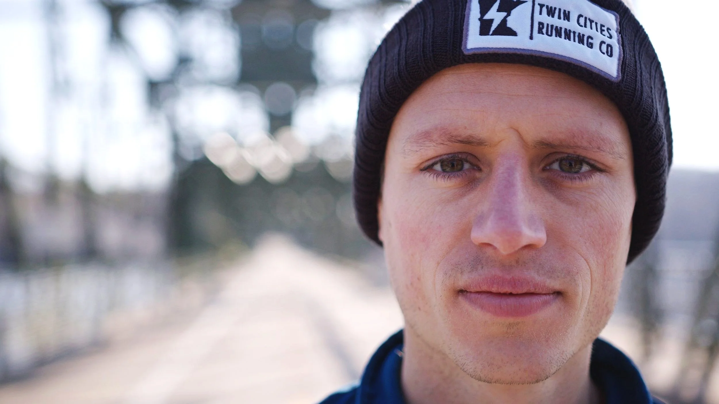 Close-up of a young man wearing a black beanie with a logo that reads 'Twin Cities Running Co,' outdoors with blurred background.