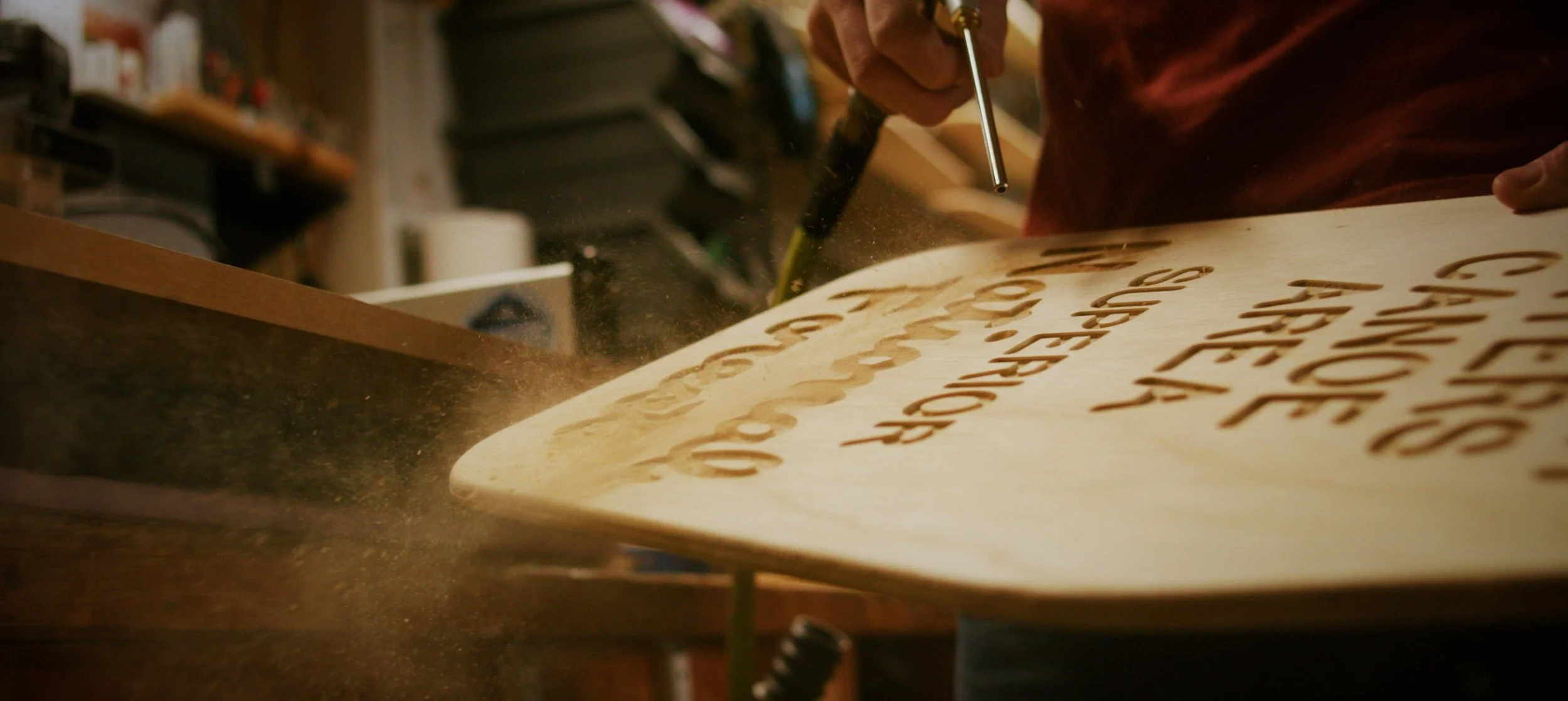 A person is using a rotary tool to carve words into a piece of wood, creating laser-cut style text on the wood surface.