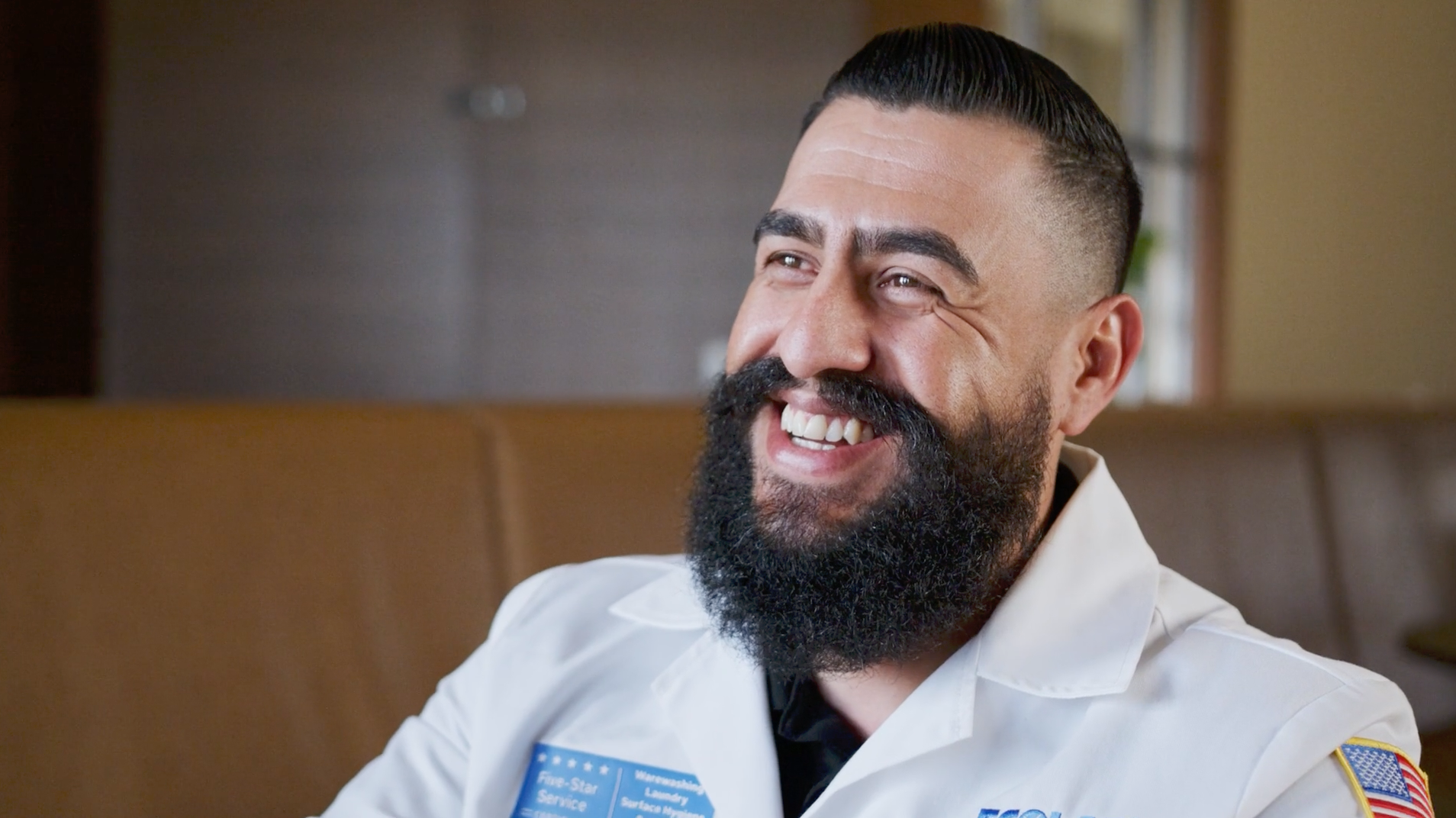 A man with a dark beard and mustache, smiling, wearing a white uniform with a name tag and an American flag patch on his arm, sitting in a restaurant or cafe.