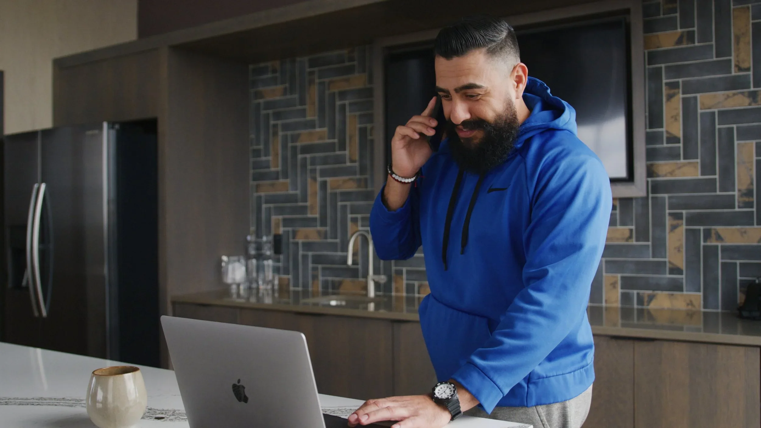 A man with a beard wearing a blue hoodie talking on his cellphone while working on a silver Apple MacBook in a modern kitchen.