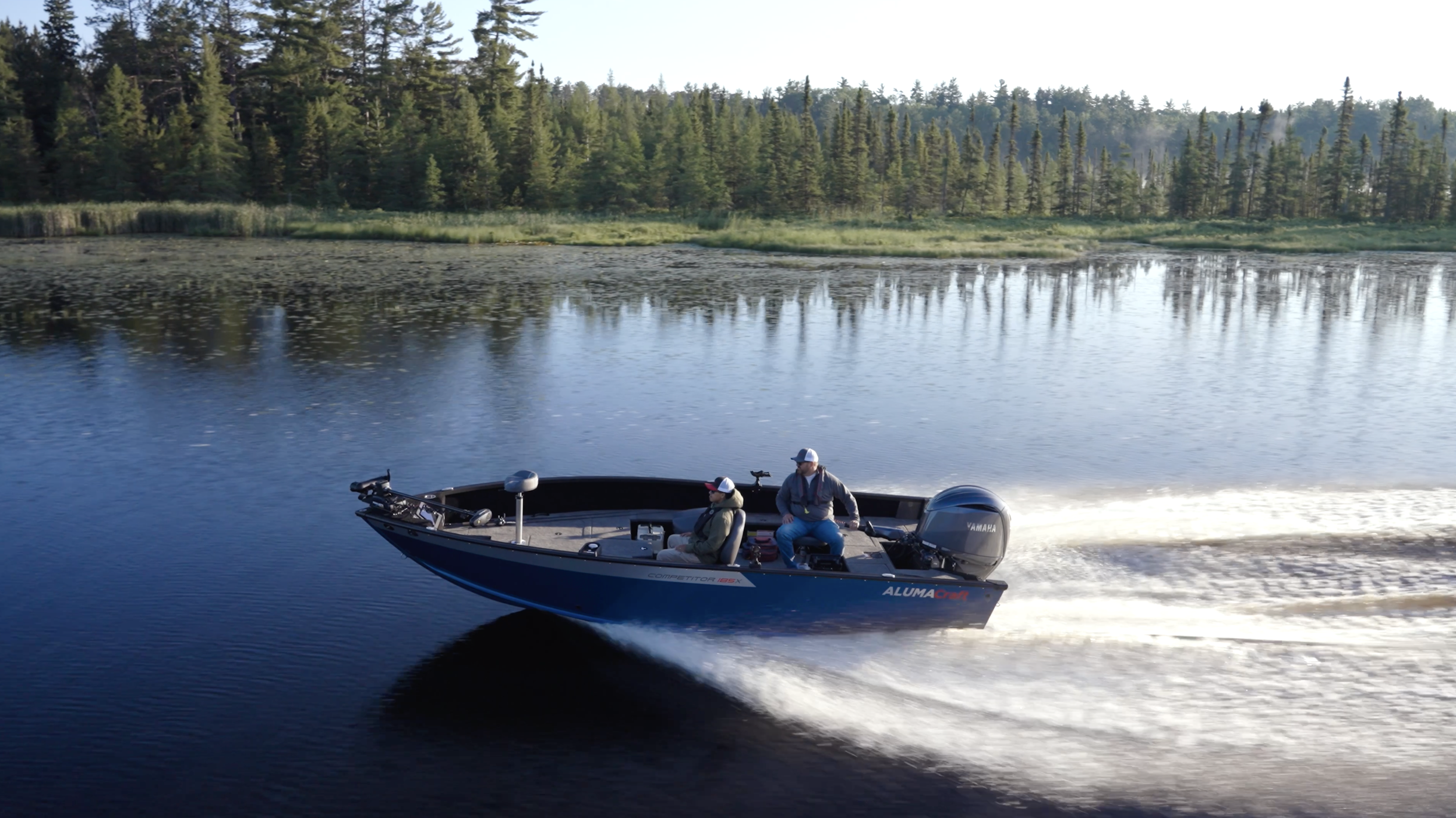 A boat with two people cruising on a lake with a tree-lined shoreline in the background.