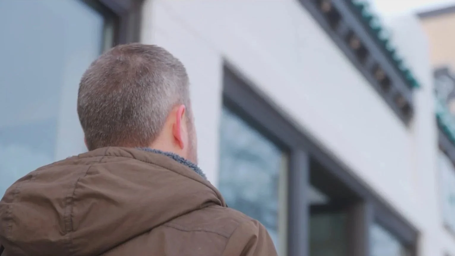 Man with short gray hair and a brown jacket looking at a modern building with glass windows.