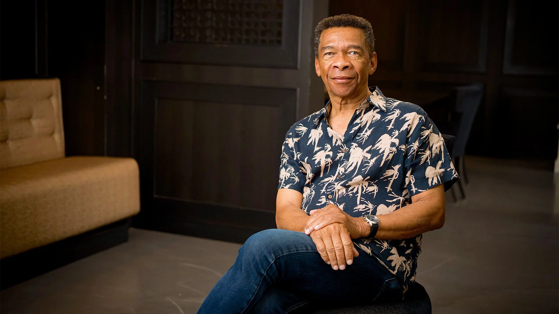 A middle-aged man with short curly hair and a tan complexion, wearing a navy blue short-sleeved shirt with a beige palm tree pattern, sitting on a stool with his legs crossed in a dark, modern room with wooden paneling. The man is looking slightly to the right of the camera, with a calm expression and a slight smile.