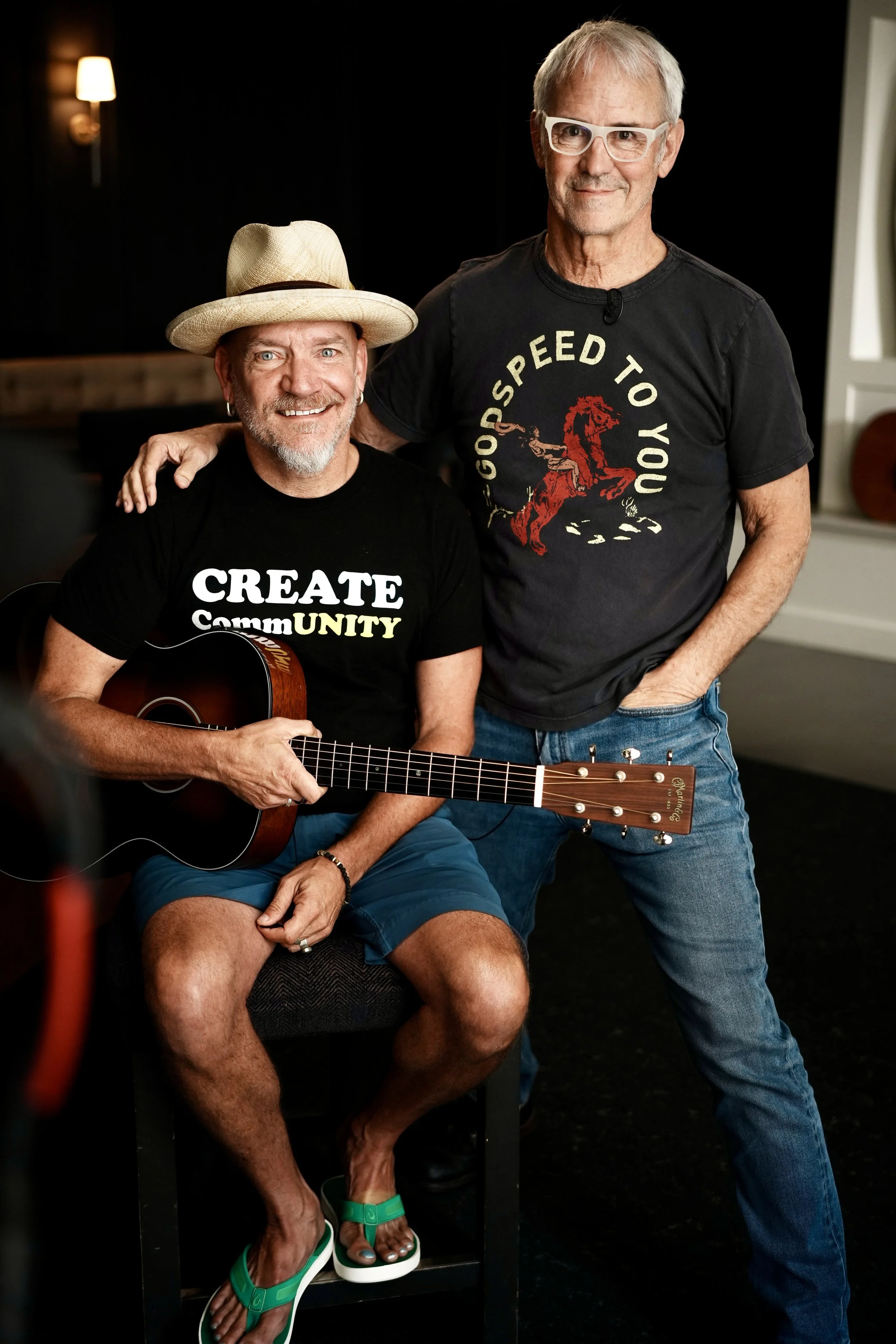 Two men in a room, one sitting with a guitar, wearing a hat, shorts, and flip-flops, smiling. The other standing, wearing glasses and a black T-shirt, posing with his hand on the seated man's shoulder.