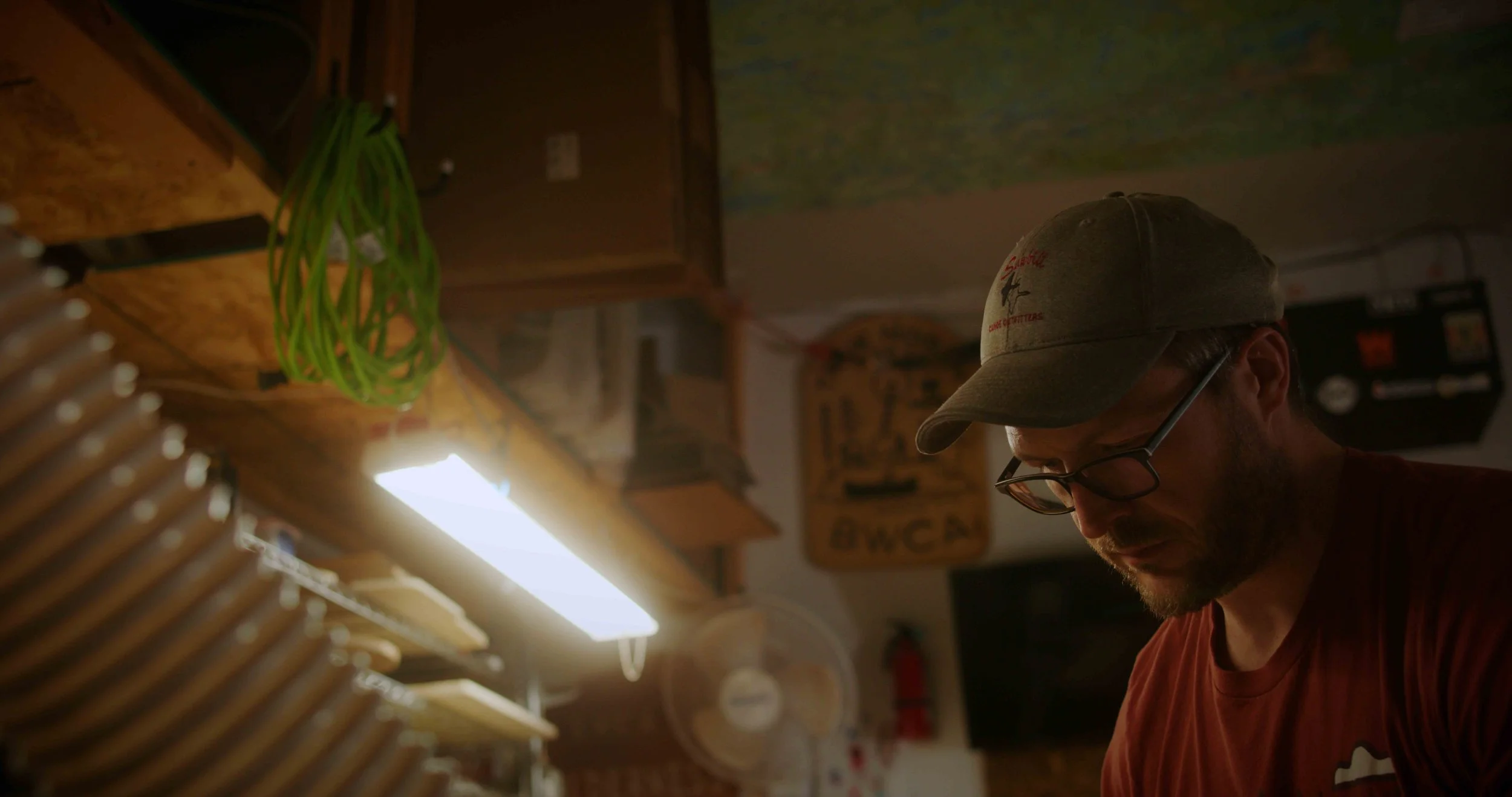 A man with glasses and a baseball cap looking down in a wood-paneled room with hanging green wires and various signs.