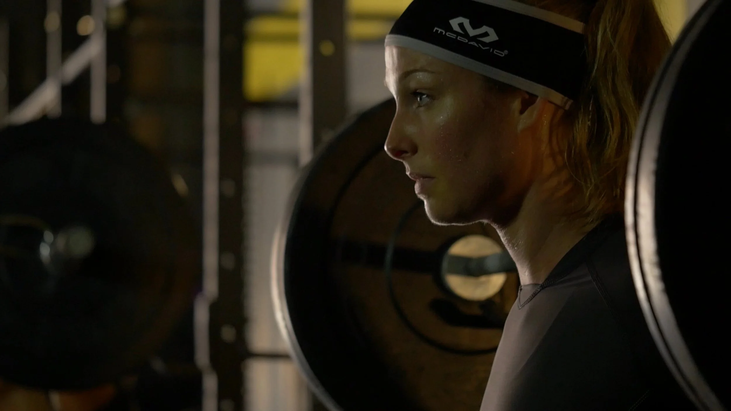 A woman with sweat on her face wearing a black sports shirt and headband, standing in a gym surrounded by weightlifting equipment.