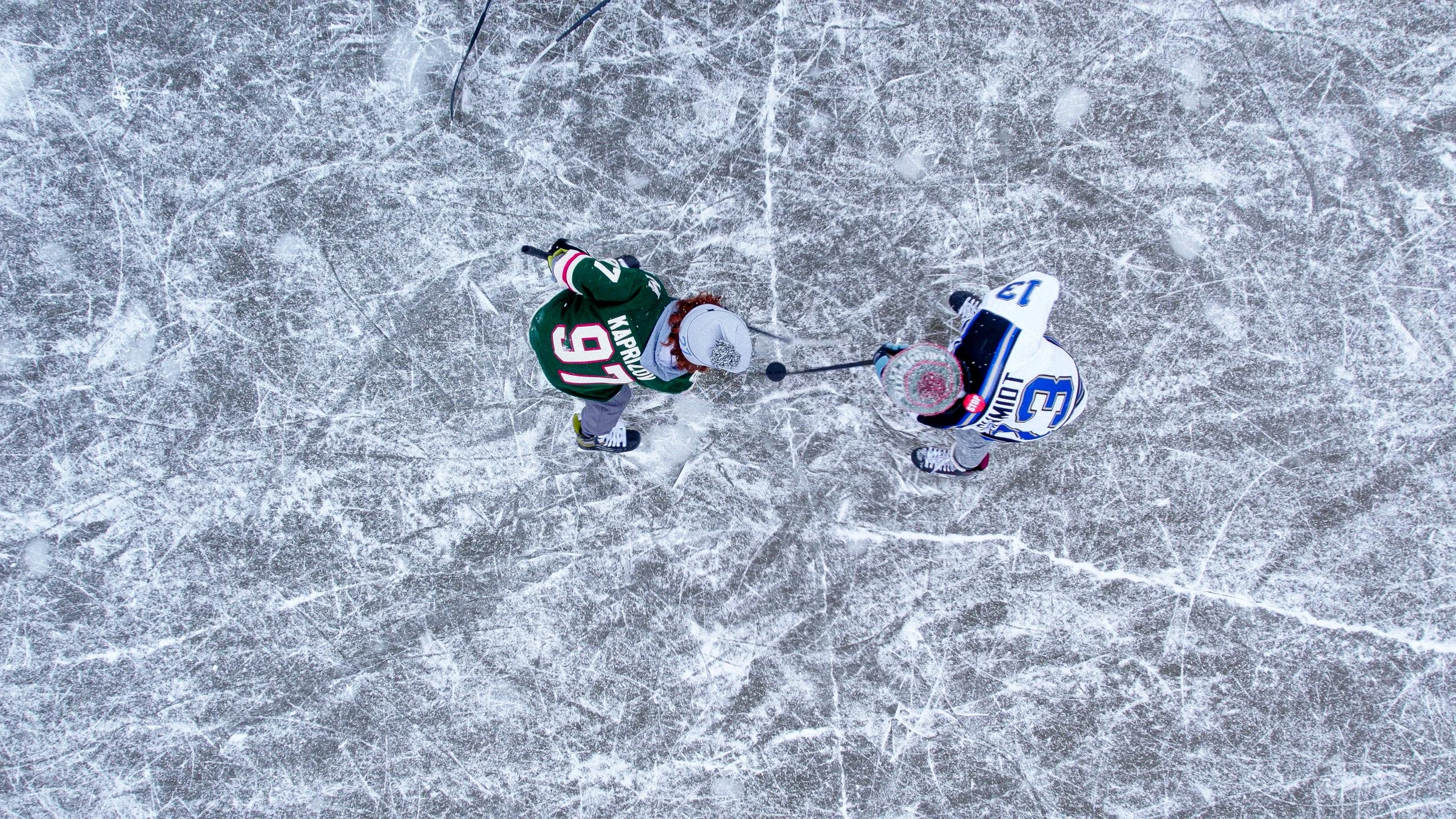 Overhead view of two hockey players on an ice rink, engaging in a faceoff, with one player in a green uniform and the other in a white uniform.