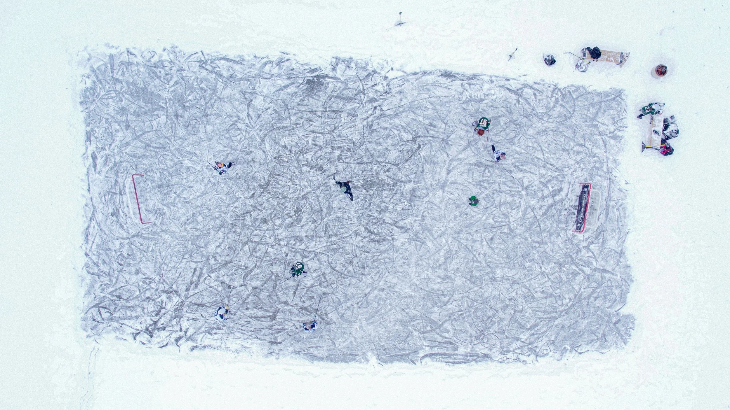Bird's-eye view of a snowy hockey rink with players, goals, and scattered equipment.