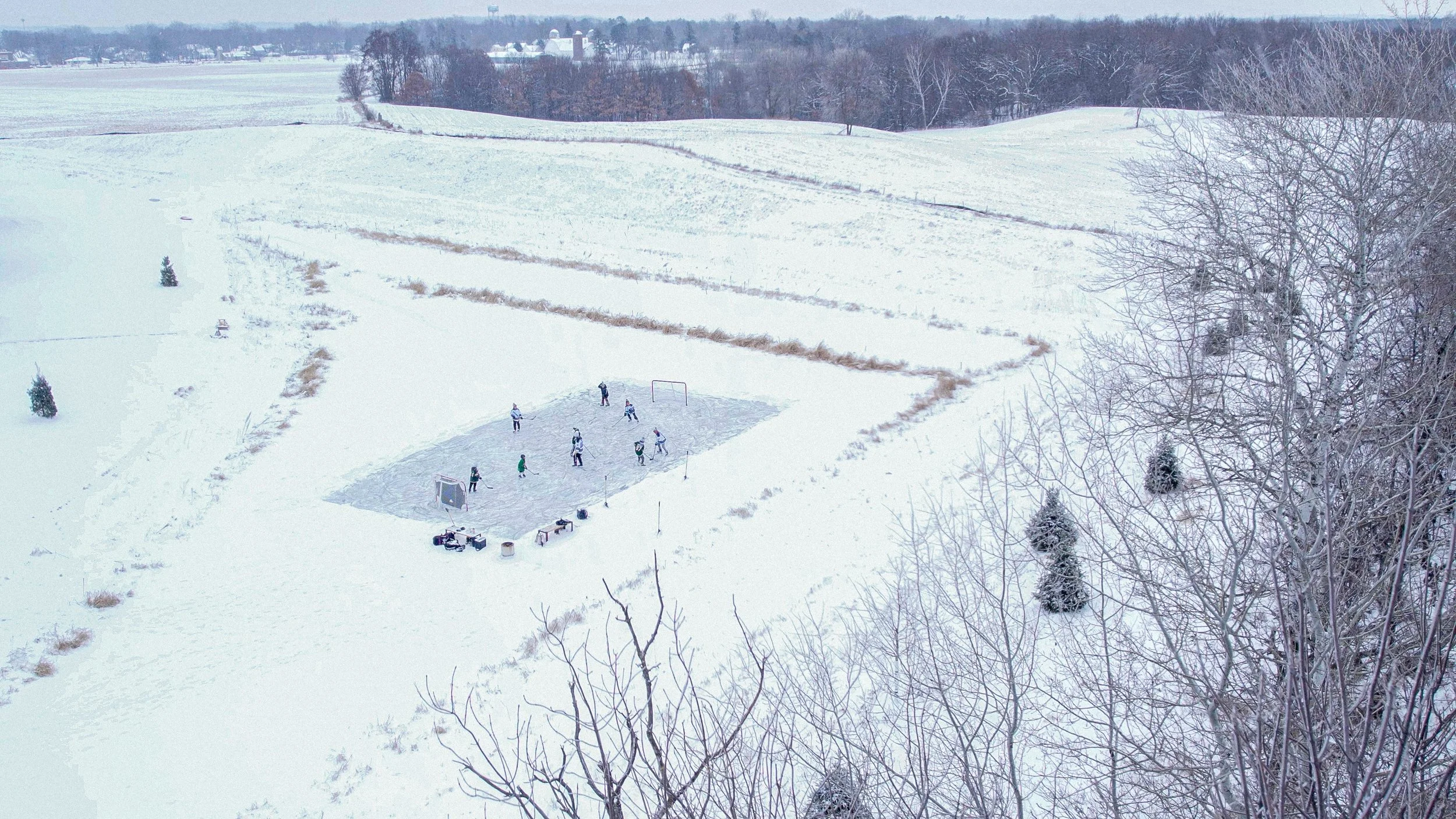 A snowy outdoor scene featuring an ice hockey game on a small rink with players, surrounded by snow-covered fields and trees, with a distant farmhouse or barn visible on the horizon.