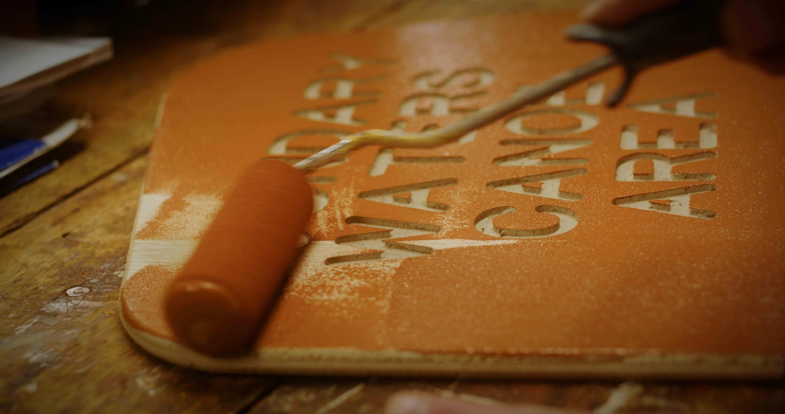Paint roller applying orange paint onto a sign with cut-out letters.