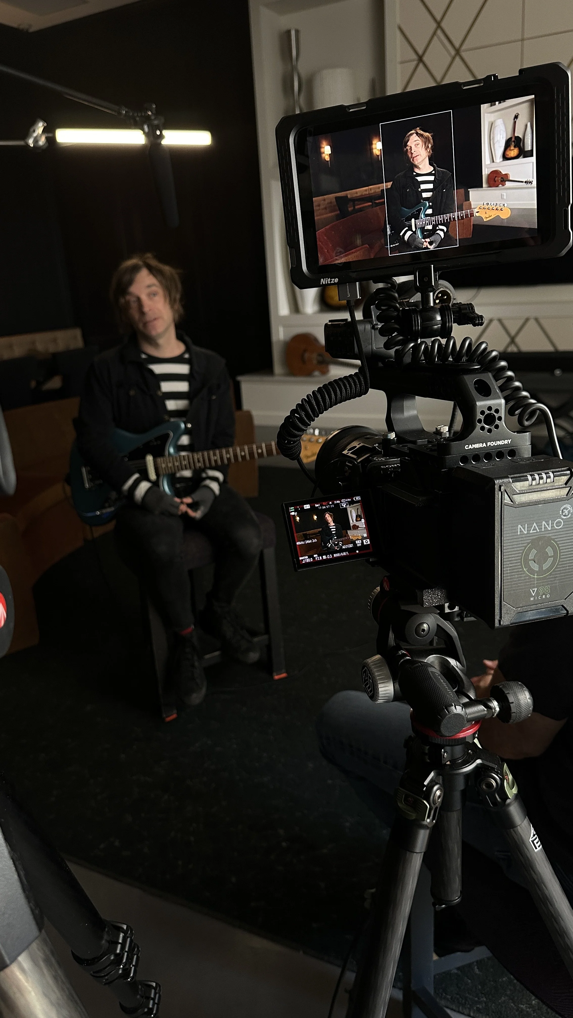Male musician filming a music video in a studio, sitting on a chair with an electric guitar, surrounded by camera equipment and lighting.