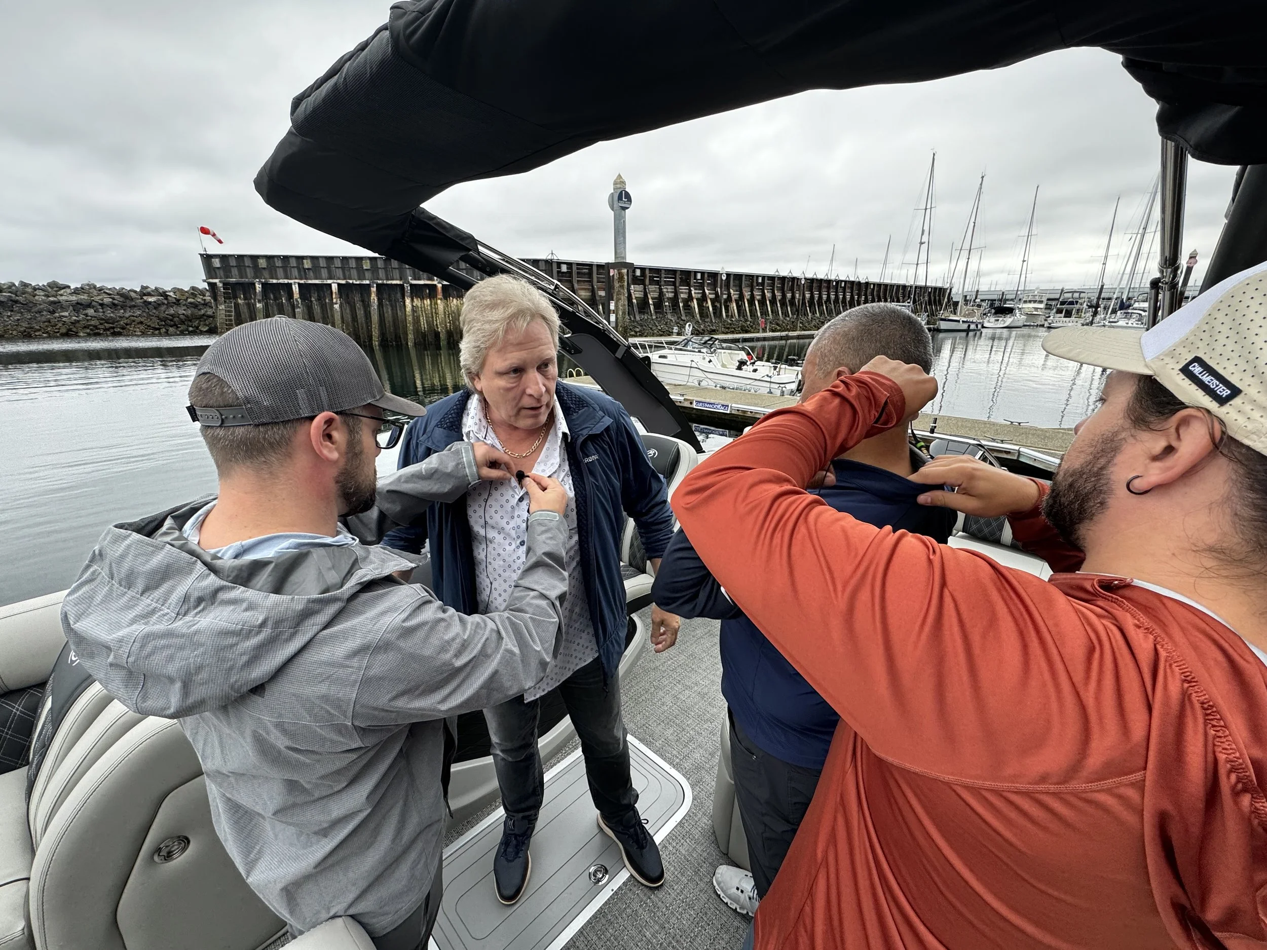A group of four men on a boat in a marina, with one man adjusting a necklace on an older man, while the others look on. The marina has numerous sailboats anchored and a stone pier in the background, under cloudy skies.