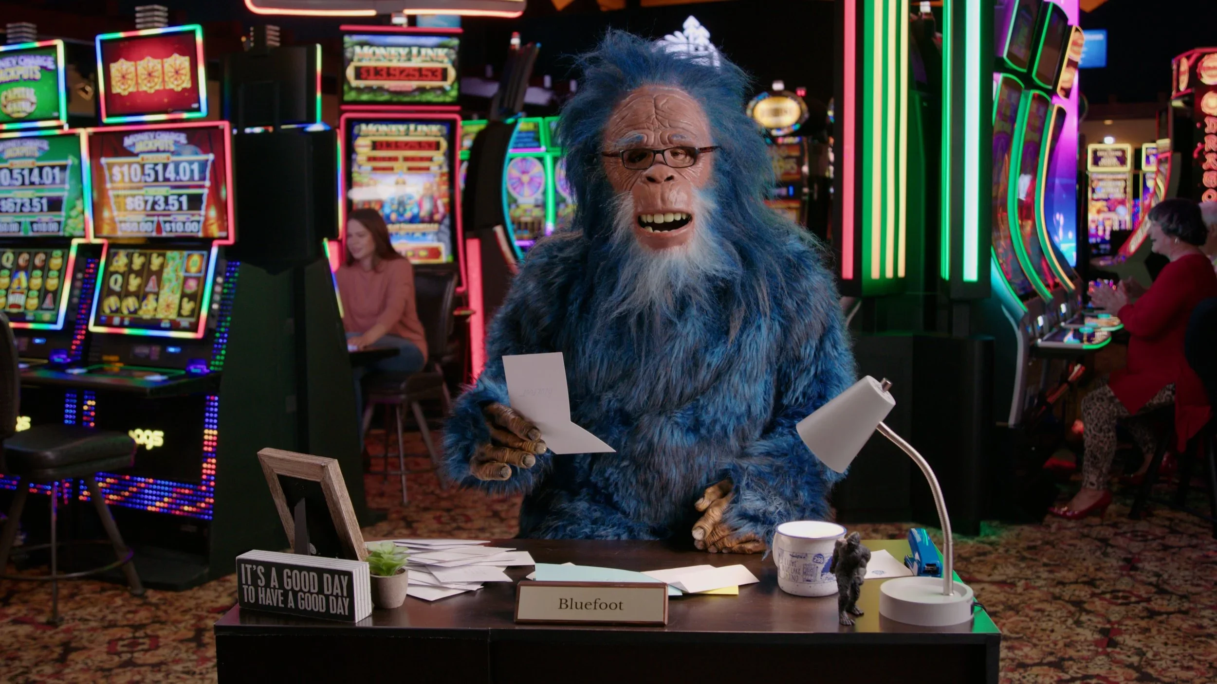 A creature with a humanoid body and a furry, blue ape-like face wearing glasses, sitting at a desk in a casino filled with brightly lit slot machines. The creature is holding a piece of paper, with a nameplate that reads 'Bluefoot', and a small black figurine on the desk. There is a lamp, a mug, a sign that says 'It's a good day to have a good day,' and papers on the desk.