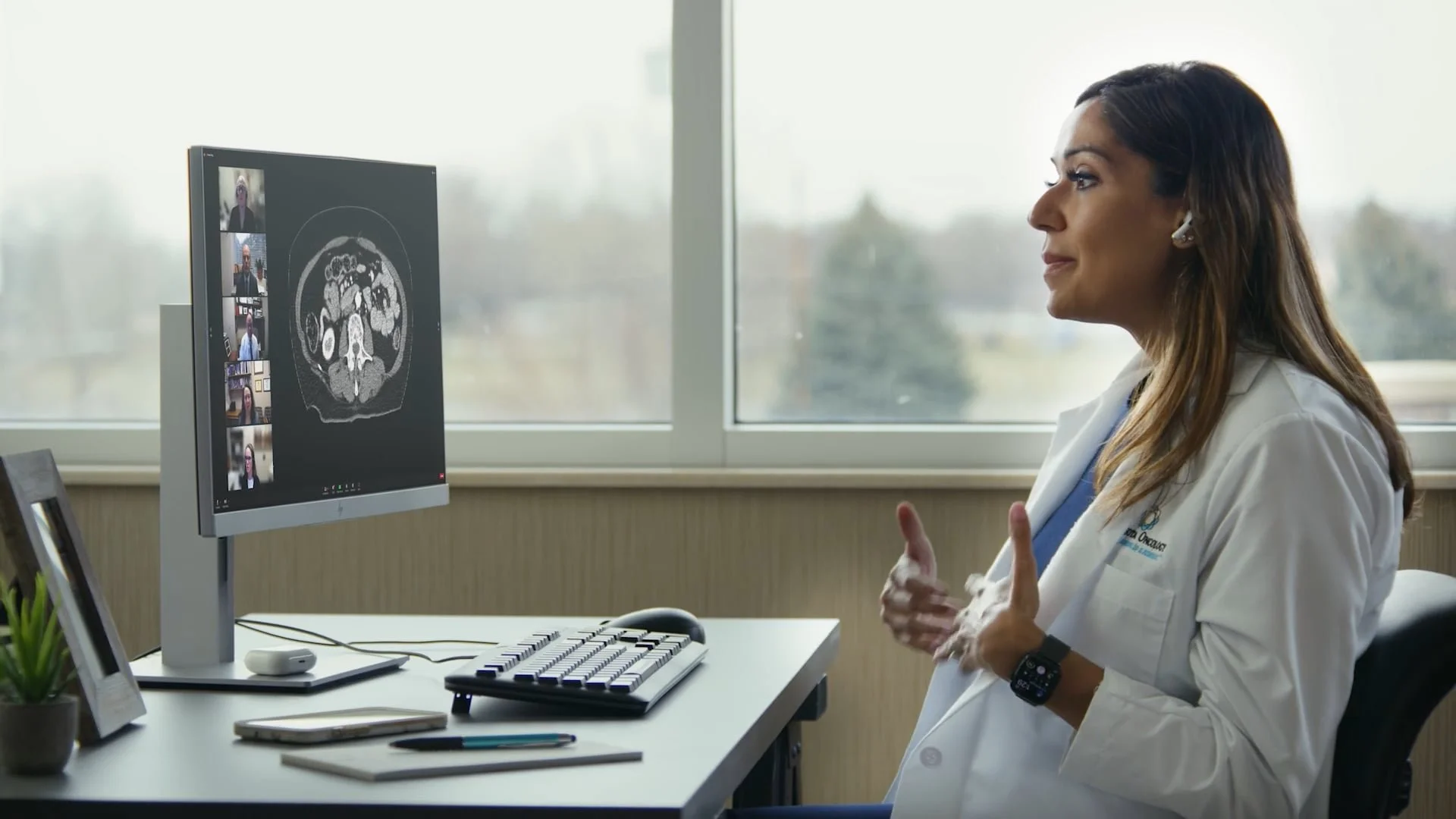 A female doctor in a white lab coat sitting at her desk in an office, engaging in a virtual meeting on her computer with a brain scan image displayed.