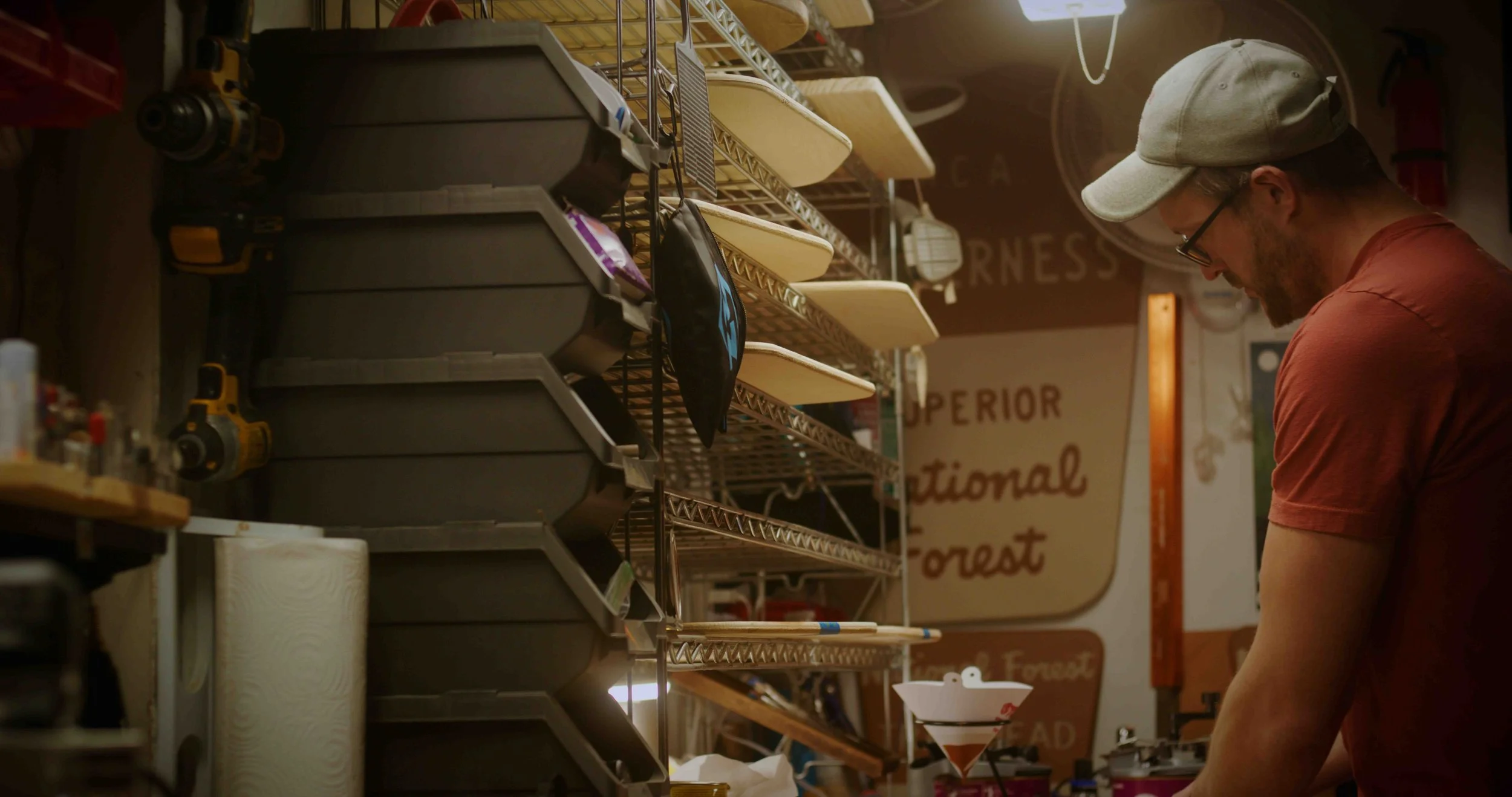 A man with a beard, glasses, a gray cap, and a red t-shirt looks down in a workshop or kitchen with metal shelves, containers, and tools around him, and a sign on the wall in the background.