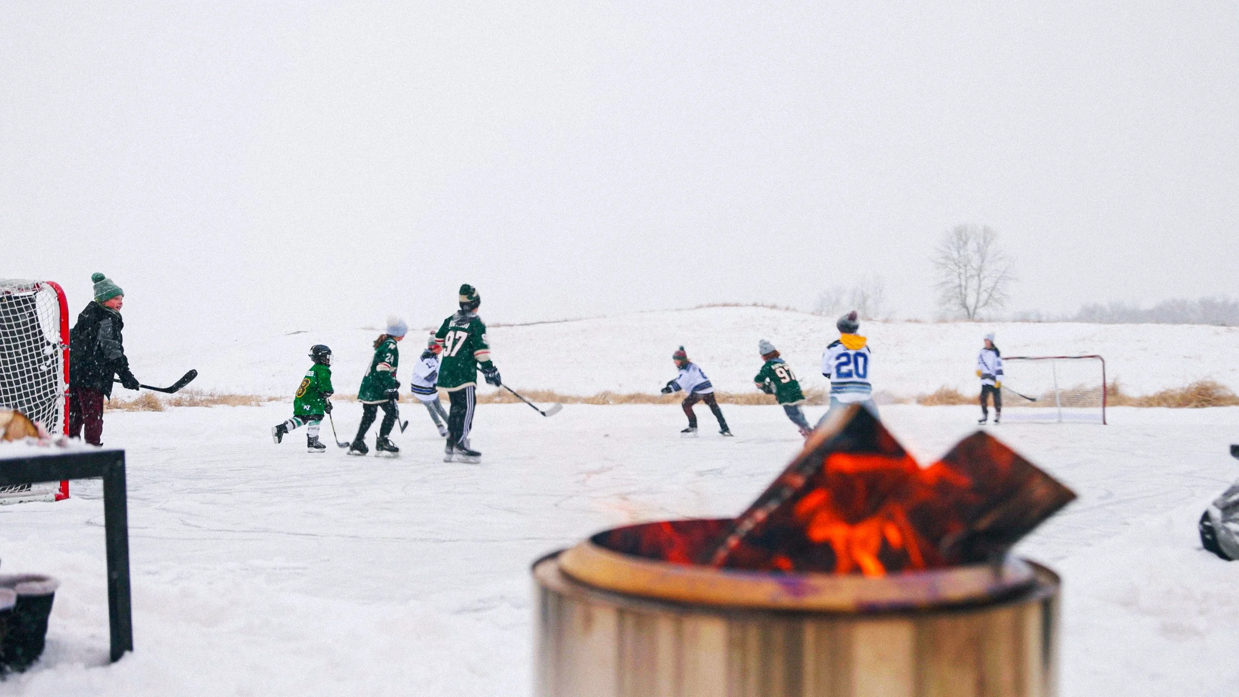 Children and adults playing hockey on an outdoor snow-covered rink with a fire in the foreground and leafless trees in the background.
