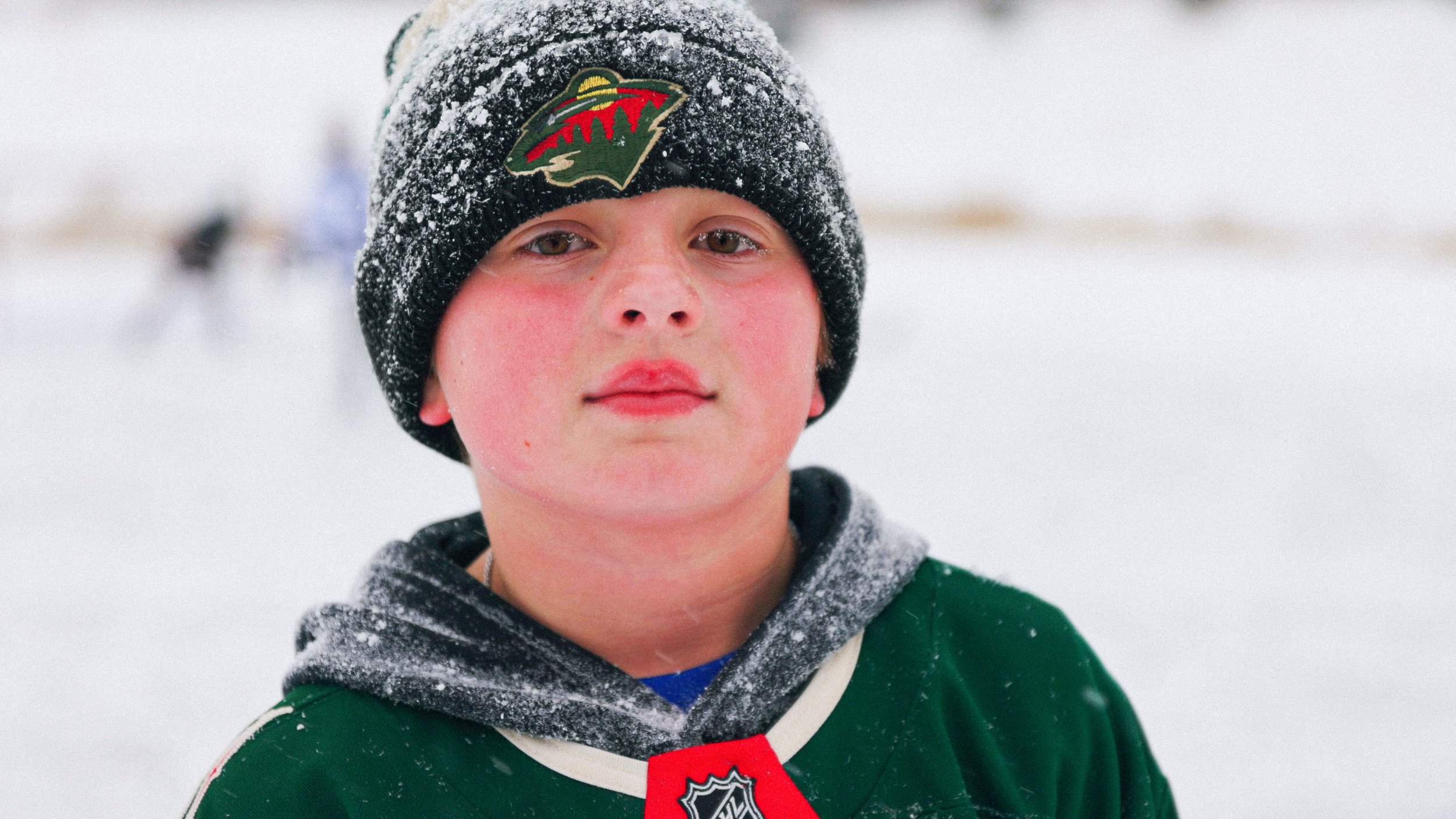 Close-up of a young boy in winter clothing, wearing a black Minnesota Wild hockey beanie with snow on it, standing in a snowy outdoor setting with a blurred background.