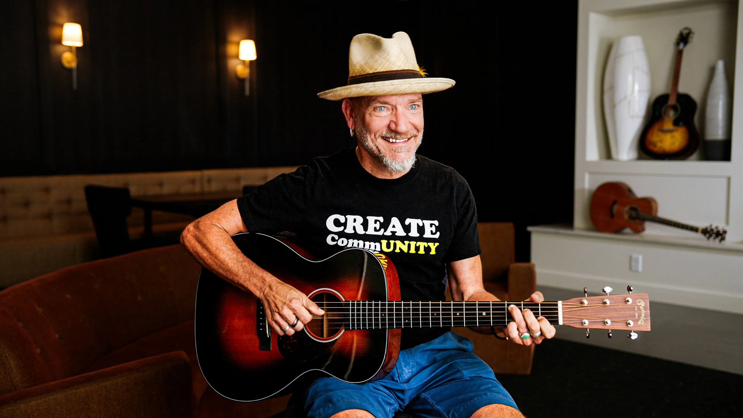 A smiling man with a beard and blue eyes wearing a straw hat, black t-shirt with 'CREATE community' written on it, and blue shorts, playing an acoustic guitar in a cozy room with dark walls, wall sconces, and shelf with decorative items and additional guitars.