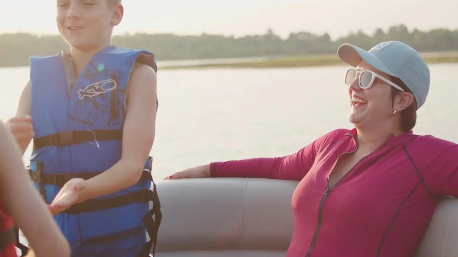 A woman wearing sunglasses and a cap, smiling and relaxing on a boat, with a young boy in a life jacket nearby, during a sunny day on a lake.