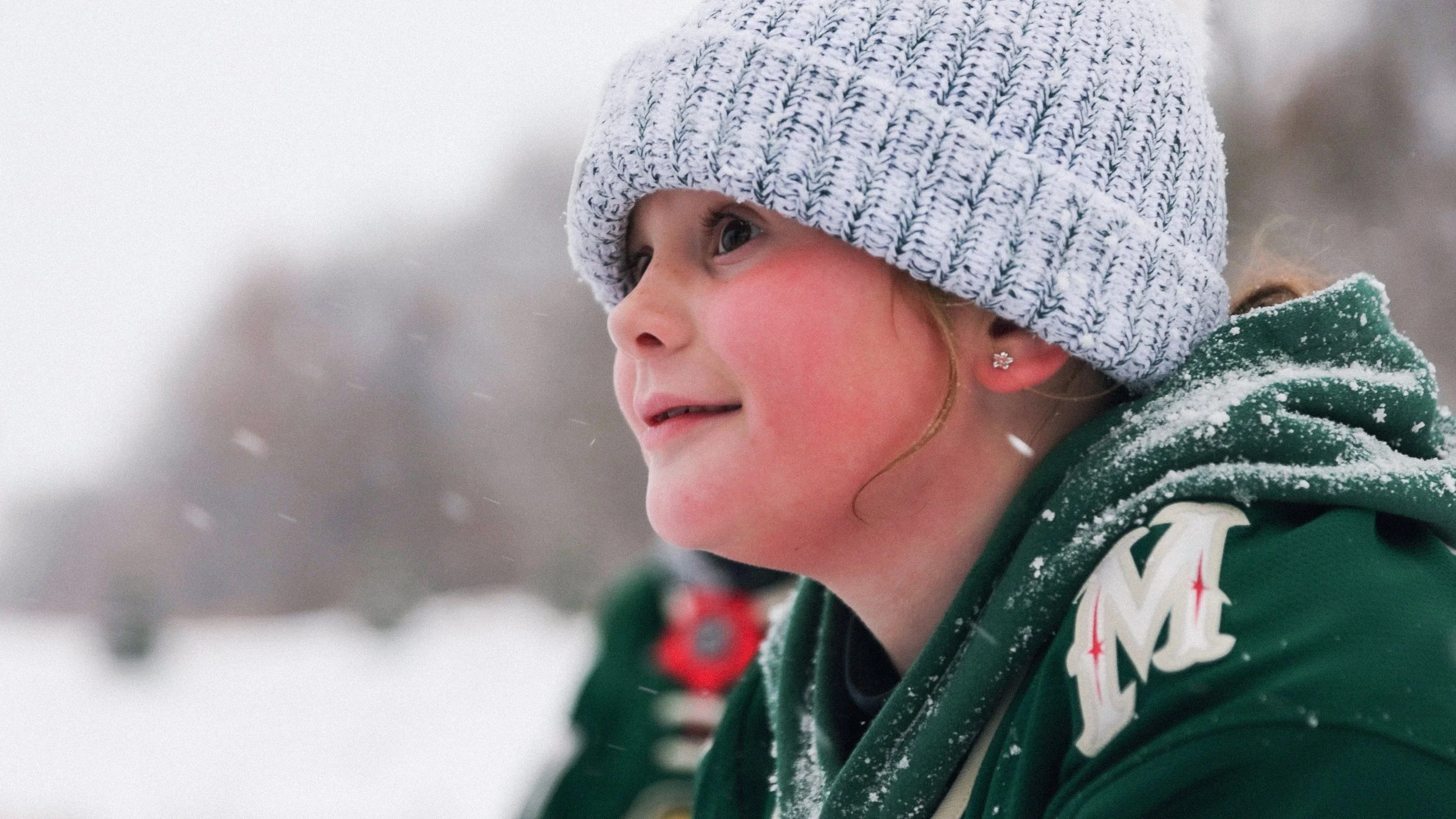 A young girl wearing a gray knit beanie and a green jacket with a logo, standing outdoors in snowy weather with snowflakes on her clothing.