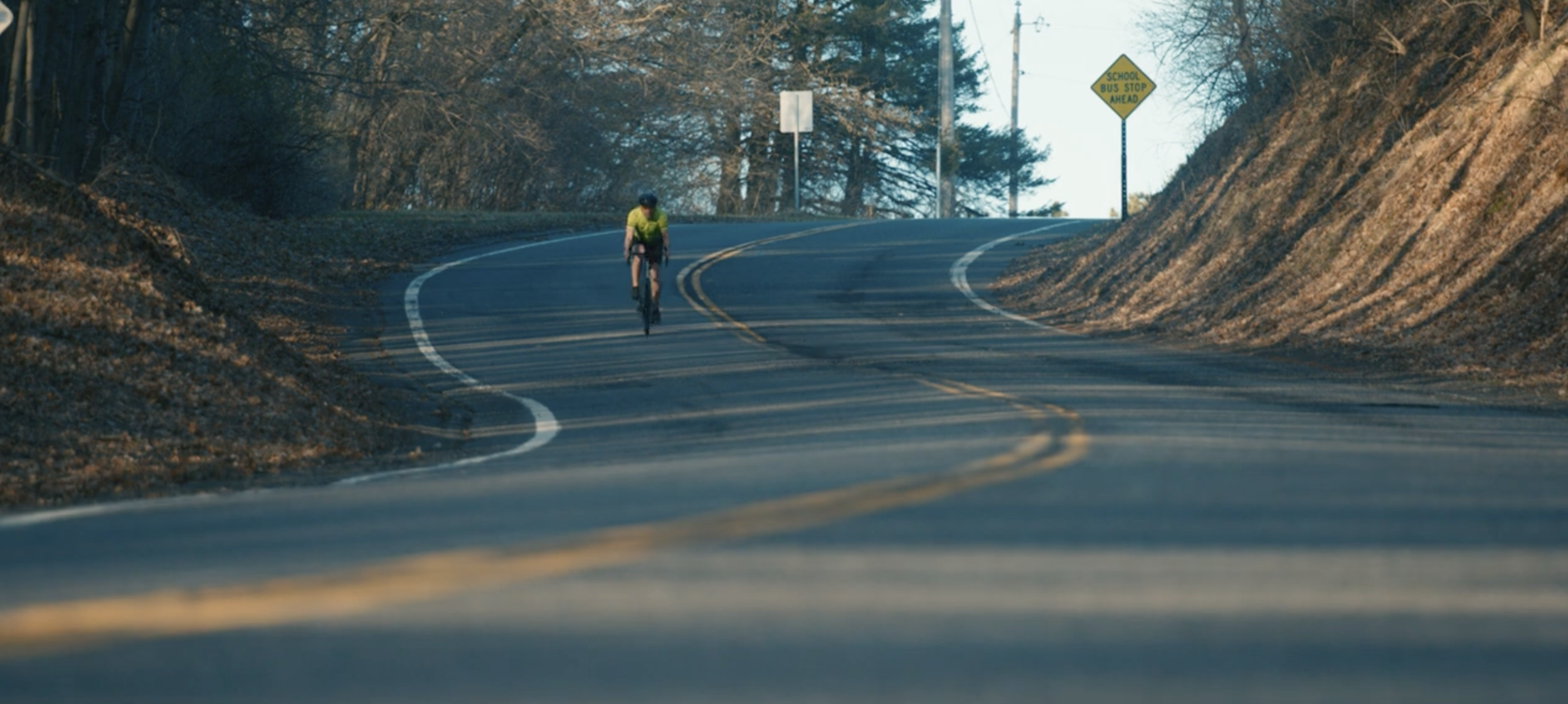 A person riding a bicycle on a winding mountain road with trees and a yellow school bus stop sign in the background.