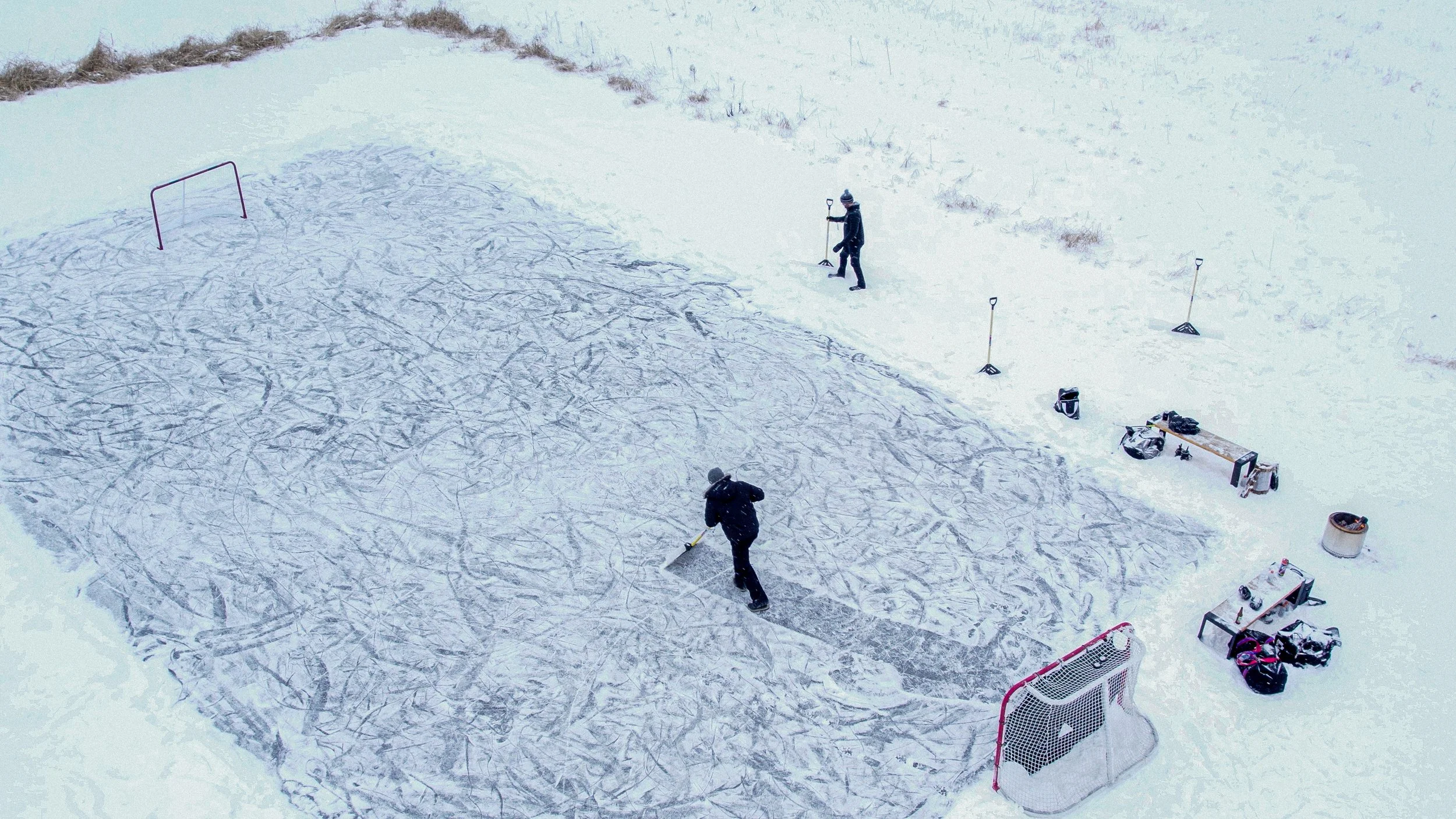 Overhead view of two people cleaning and shoveling an outdoor ice hockey rink in a snow-covered area, with hockey goals and equipment around.