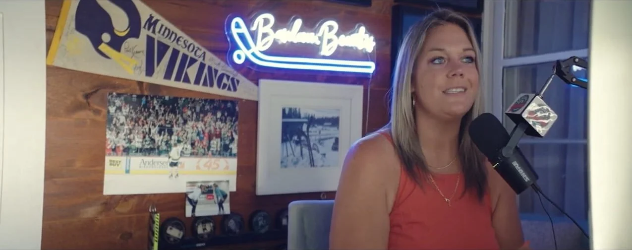 A woman is sitting in a room with wood-paneled walls, speaking into a microphone. Behind her, there are sports memorabilia including a Minnesota Vikings pennant, a framed picture, a neon sign, and a hockey-themed photograph. The room has a window with blue lighting.