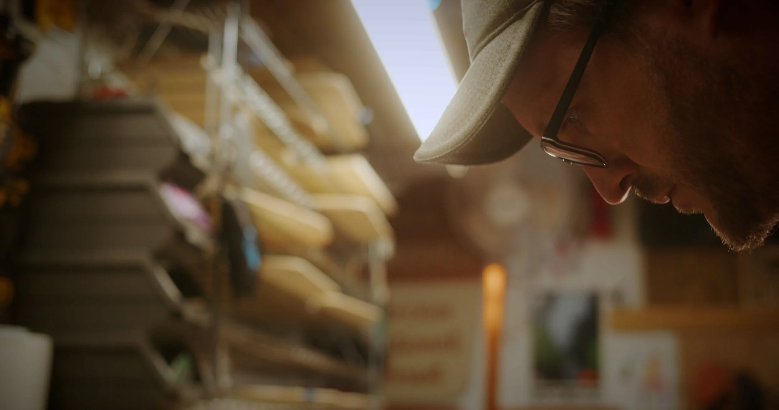 A man wearing glasses and a gray cap is closely examining items in a store, with shelves of shoes in the background.