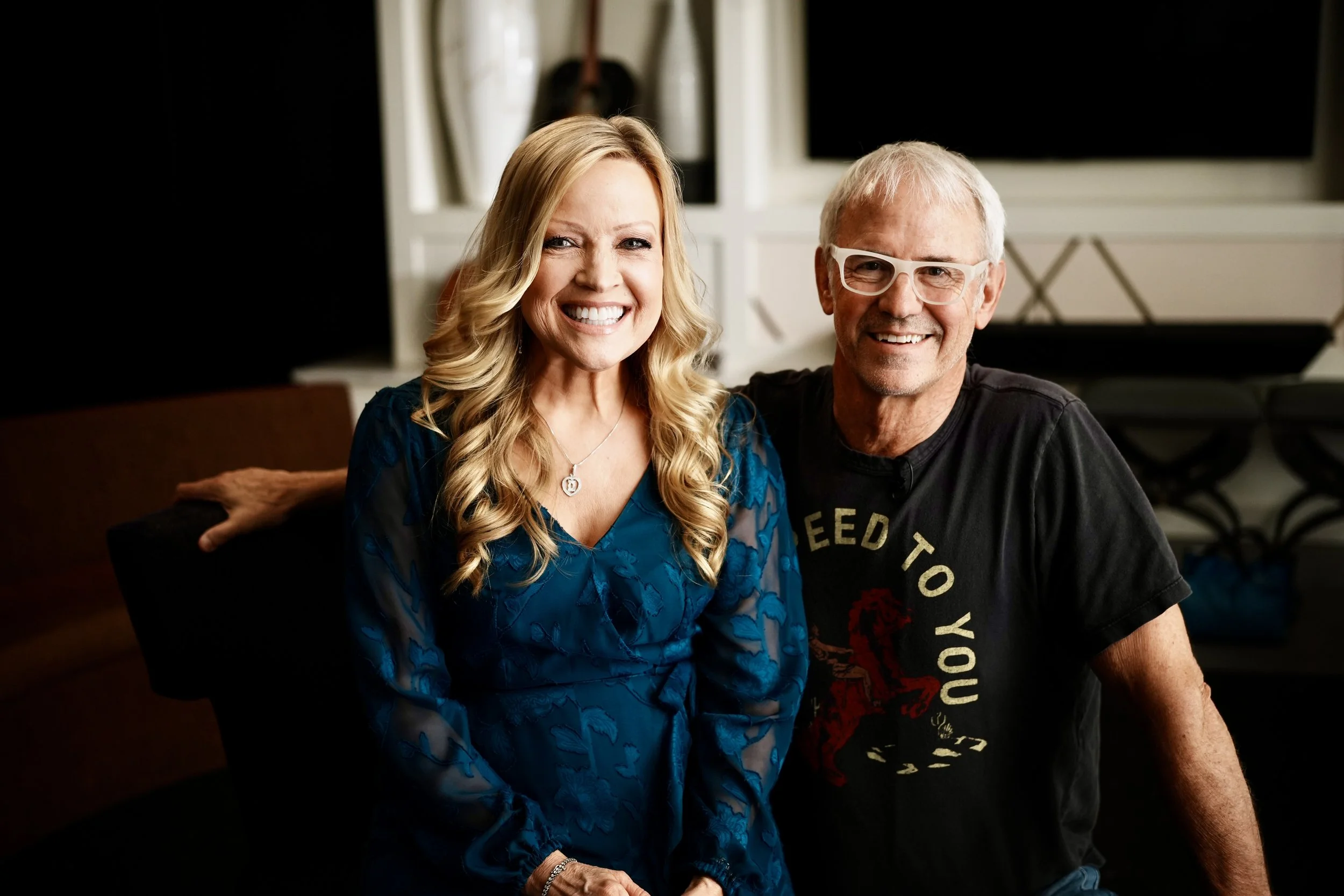 A woman with long blonde hair wearing a blue dress and a man with gray hair and glasses wearing a black t-shirt with a red T-Rex and yellow text. They are smiling and sitting in a cozy indoor setting.