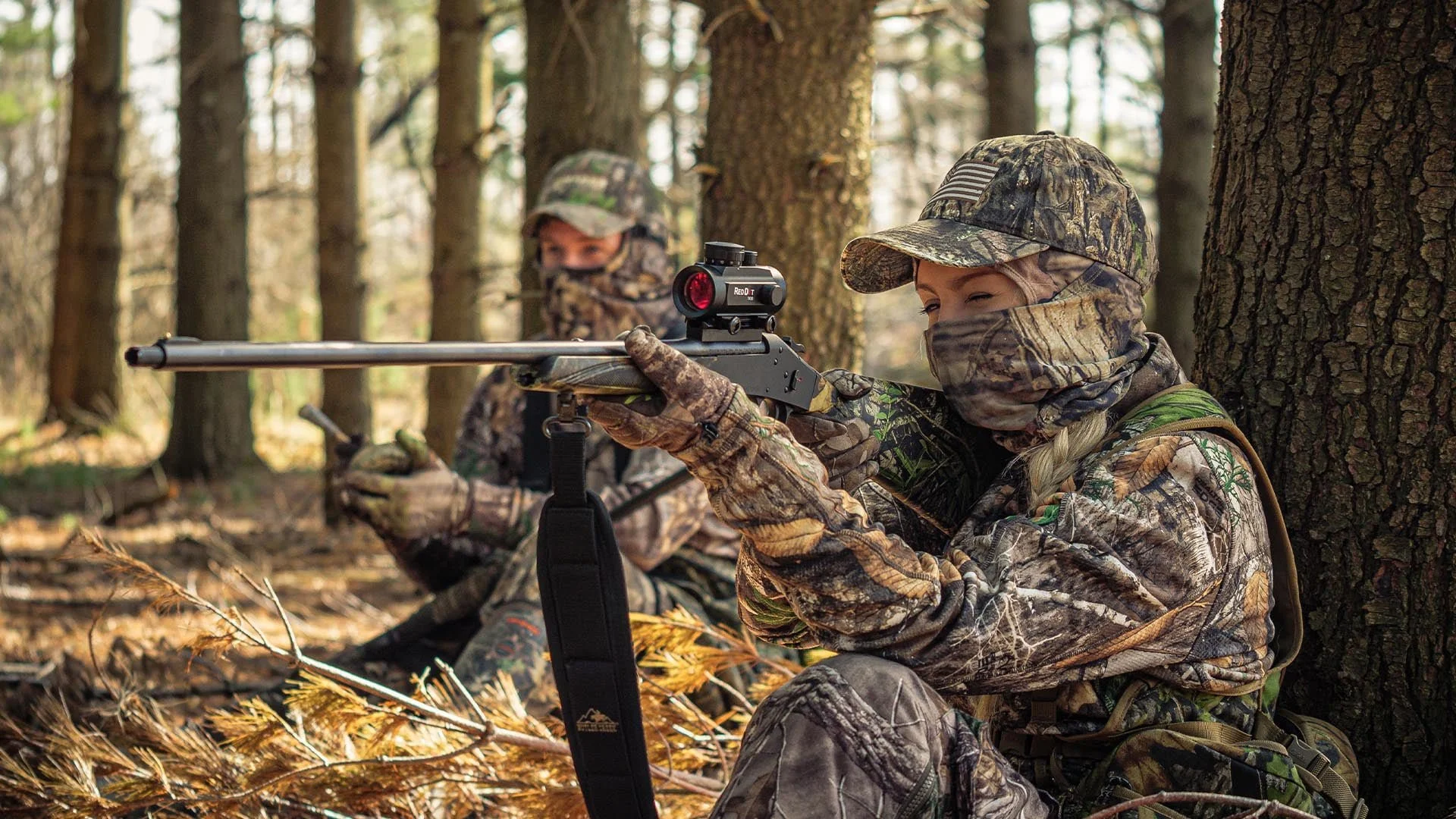 Two people dressed in camouflage hunting gear, sitting in a forest with trees and fallen leaves. One is aiming a rifle with a scope, the other is sitting behind with a face covering and a hat.