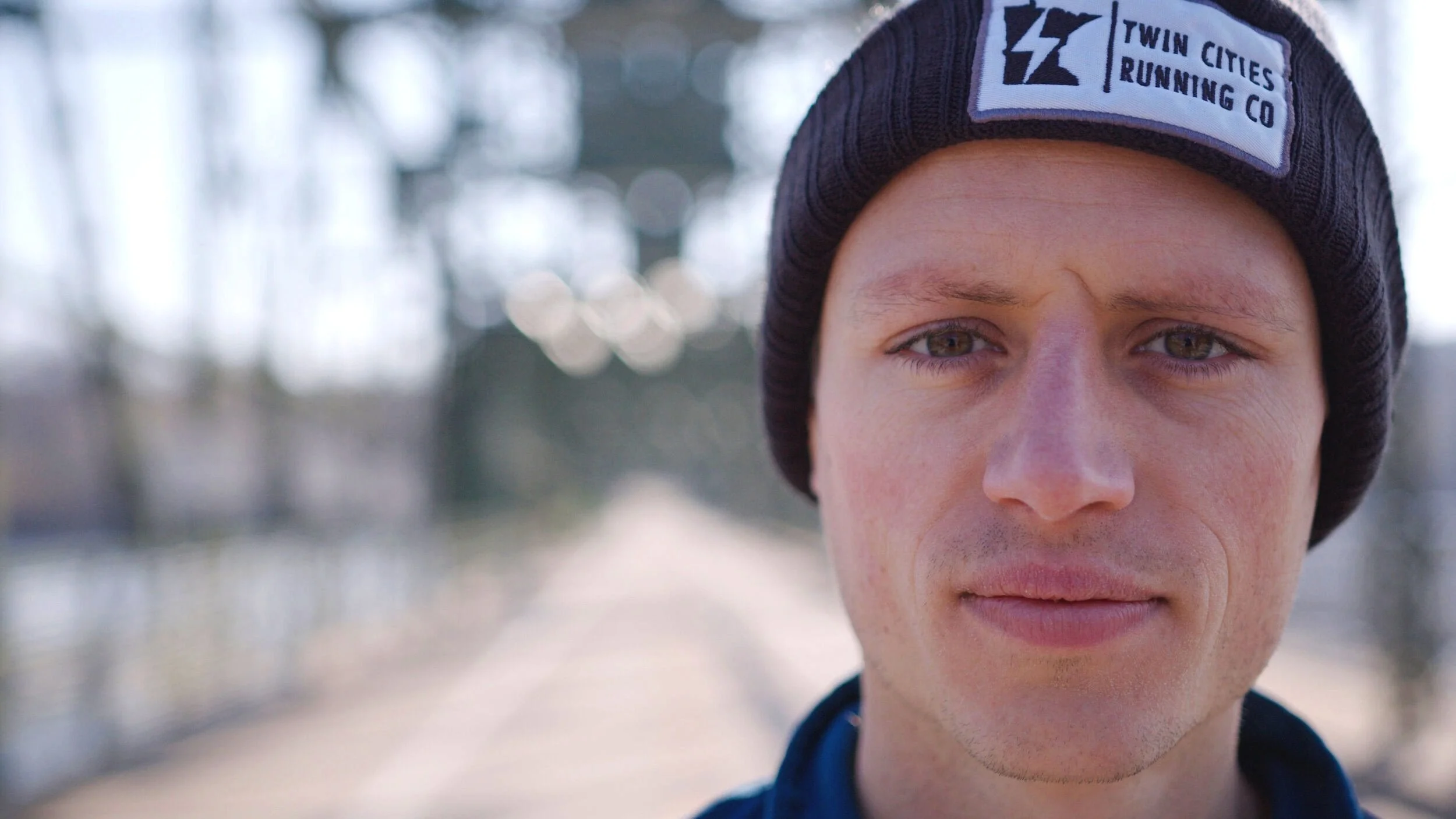 Close-up of a young man wearing a black beanie with a Twin Cities Running Co logo, outdoors in a park during daytime, with trees and a pathway blurred in the background.