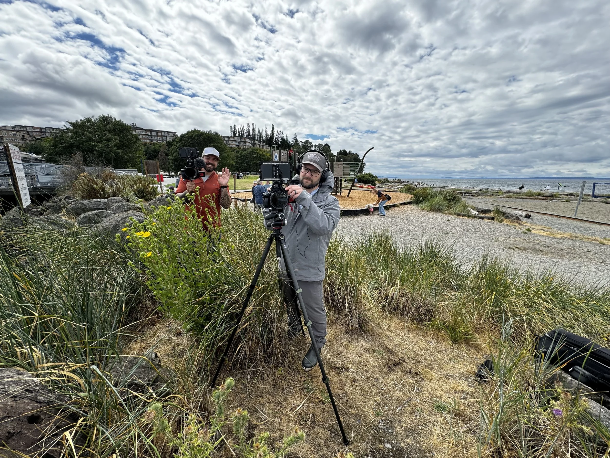 Two men film a beach scene, one operating a camera on a tripod and the other holding a handheld camera, with a cloudy sky and beach in the background.