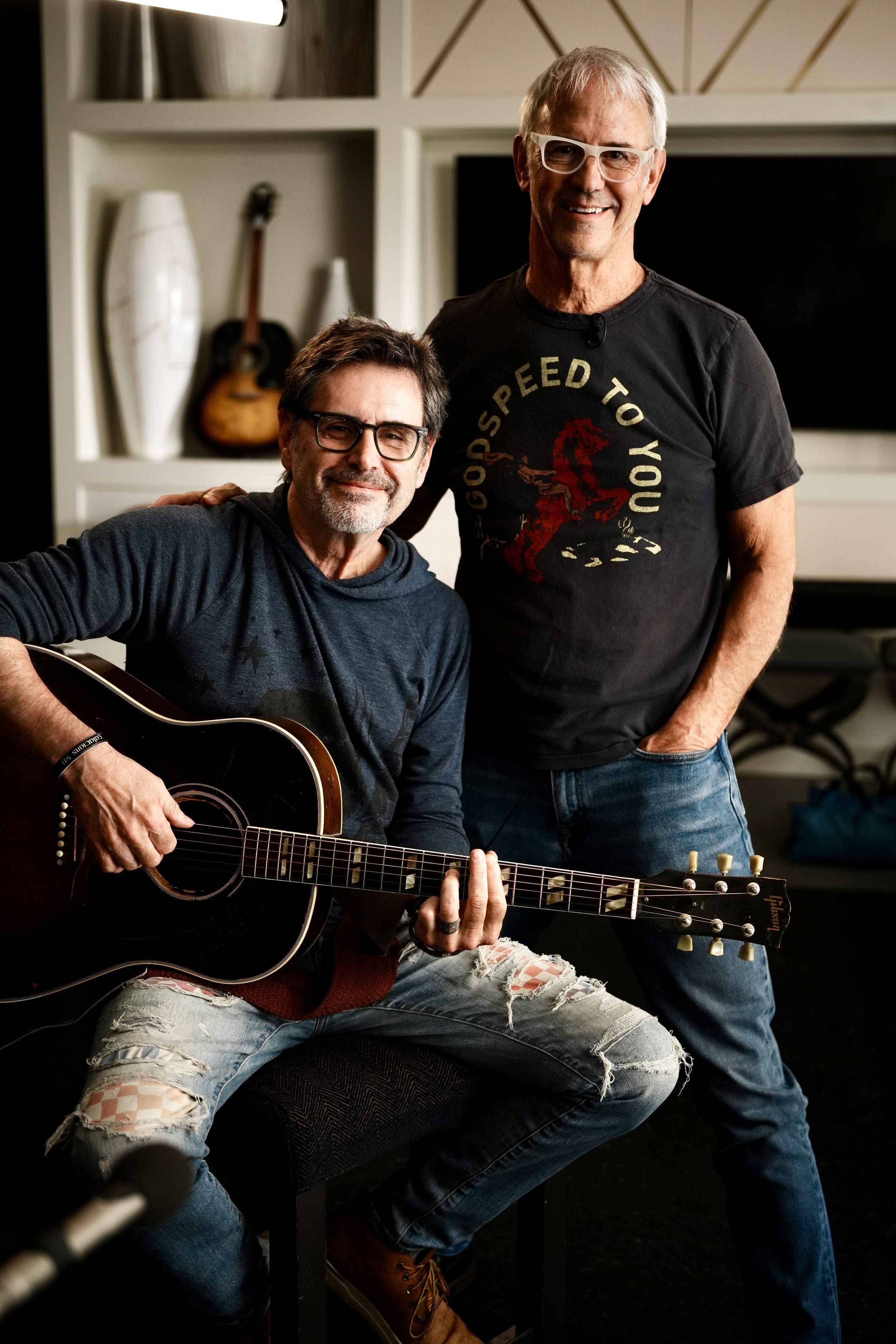 Two men smiling, one sitting with a guitar and one standing behind him, in a room with decorative objects on shelves.