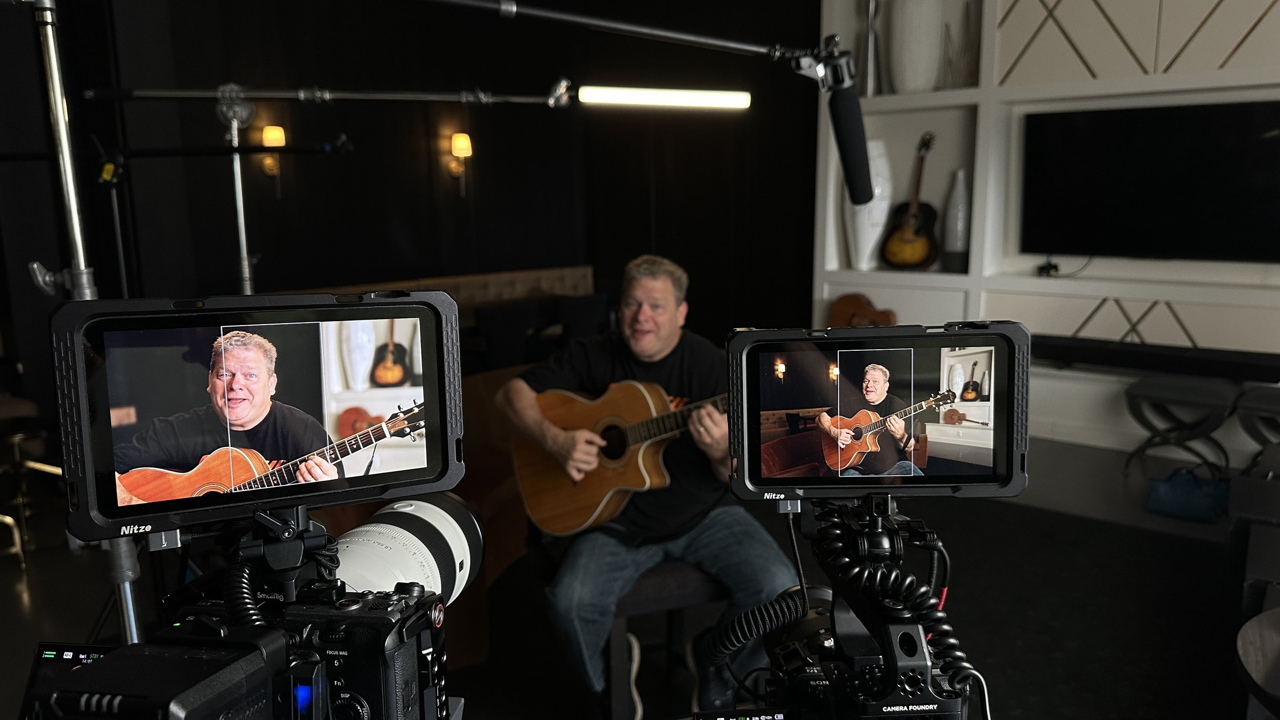 Man playing an acoustic guitar during a video recording, with two professional cameras capturing the scene in a modern indoor setting.