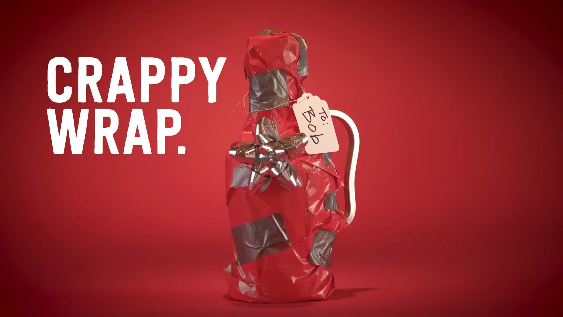 A Christmas gift wrapped in red and silver metallic paper with a silver bow, against a red background. A white tag on the gift says 'To! Dad' and there is white text that reads 'CRAPPY WRAP.'