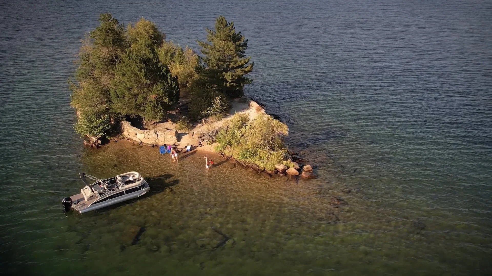 A small island with trees and rocks, with a few people on the sandy shore, a boat anchored nearby in clear green water.