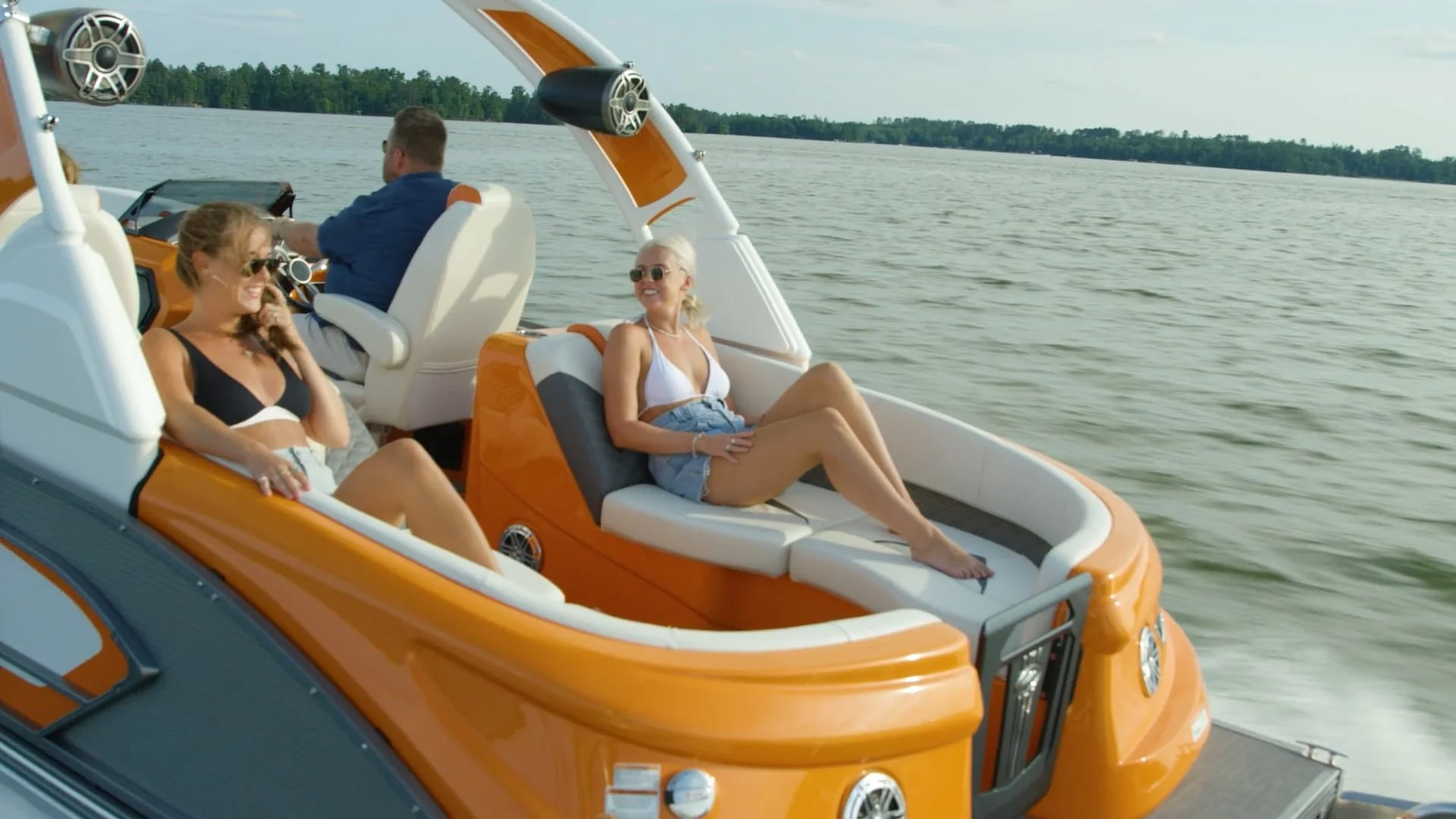 Three people on a boat with orange and white seating, enjoying a sunny day on the water. One woman in a black bikini top and shorts talking on the phone, another woman in white bikini top and shorts relaxing, and a man steering the boat.