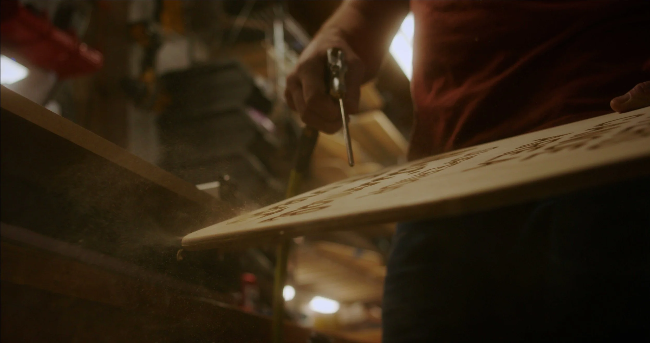 Close-up of a person using a tool on a piece of wood in a woodworking shop, with sawdust in the air.