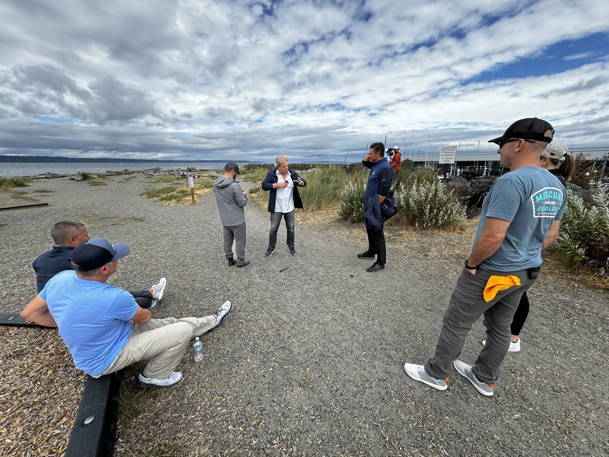 Group of people gathered outdoors on a cloudy day near a body of water, some sitting on a bench and others standing, with a dock and boats in the background.