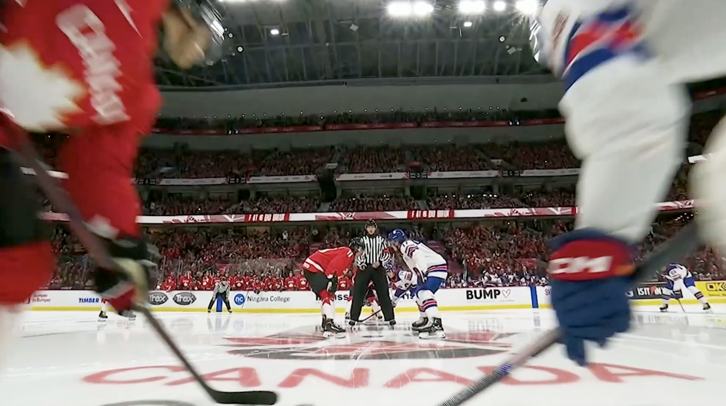 Ice hockey game with players from opposing teams about to drop the puck at center ice, surrounded by fans in an arena.
