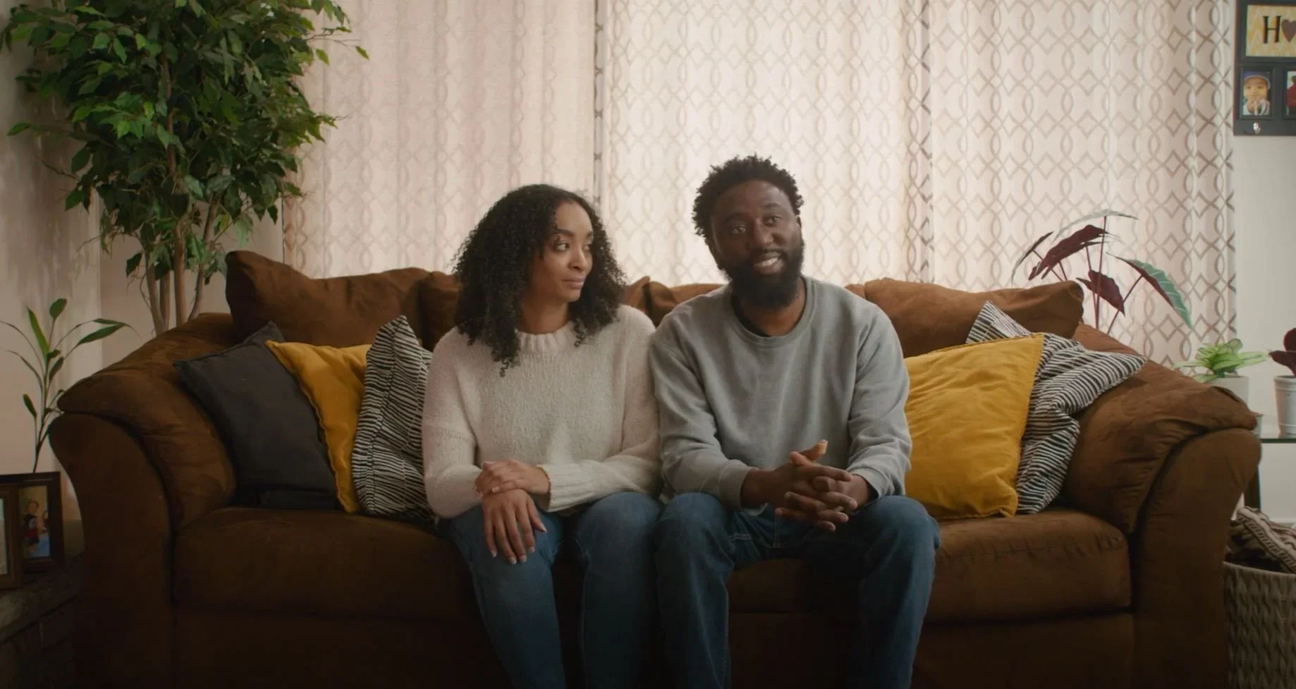 A young couple sitting on a brown sofa in a living room with beige curtains, surrounded by various plants and cushions, engaging in a conversation.