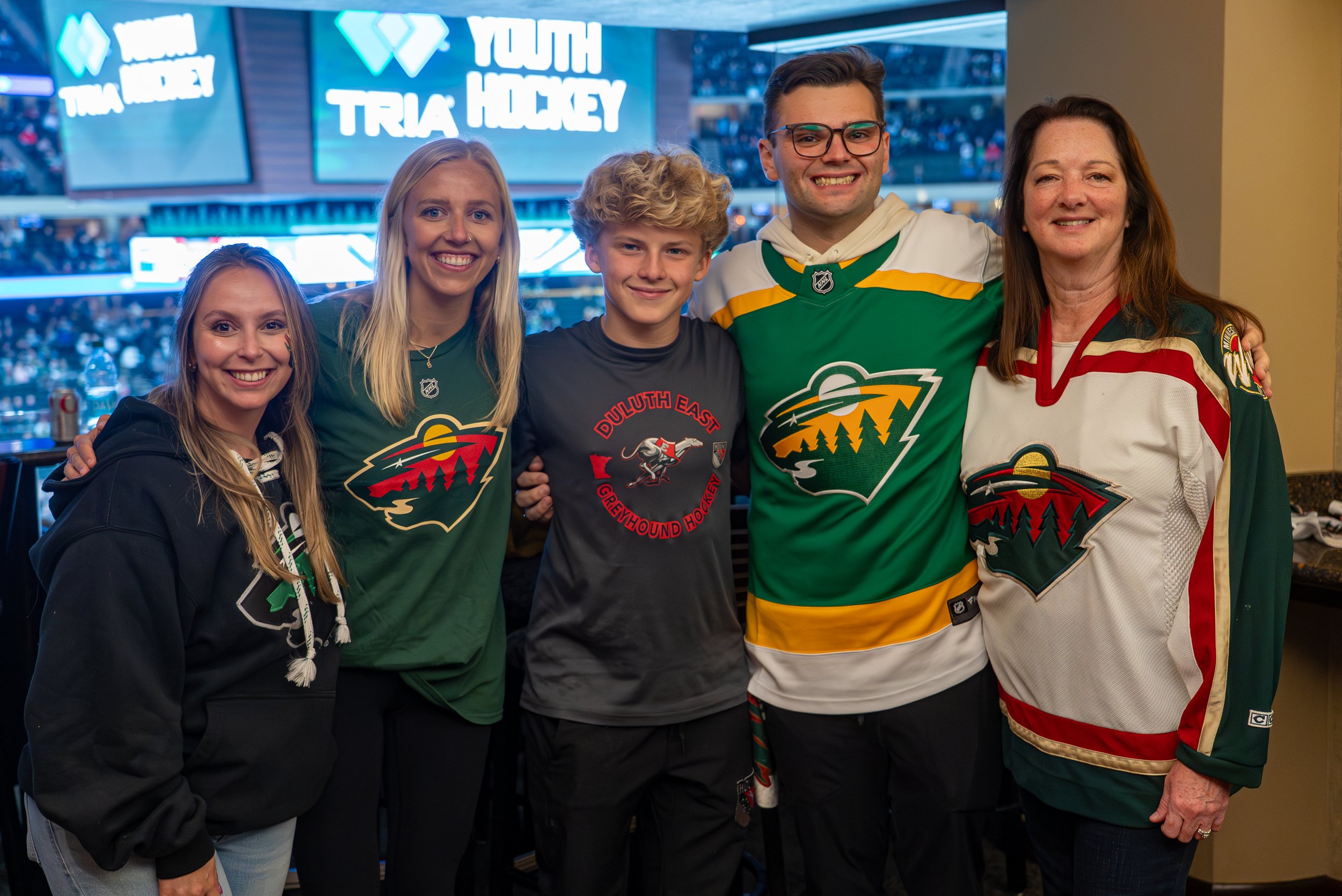 Group of six people smiling, wearing Minnesota Wild hockey jerseys, at a game with a large TV screen in the background displaying 'Youth Hockey' and 'Trivia'.