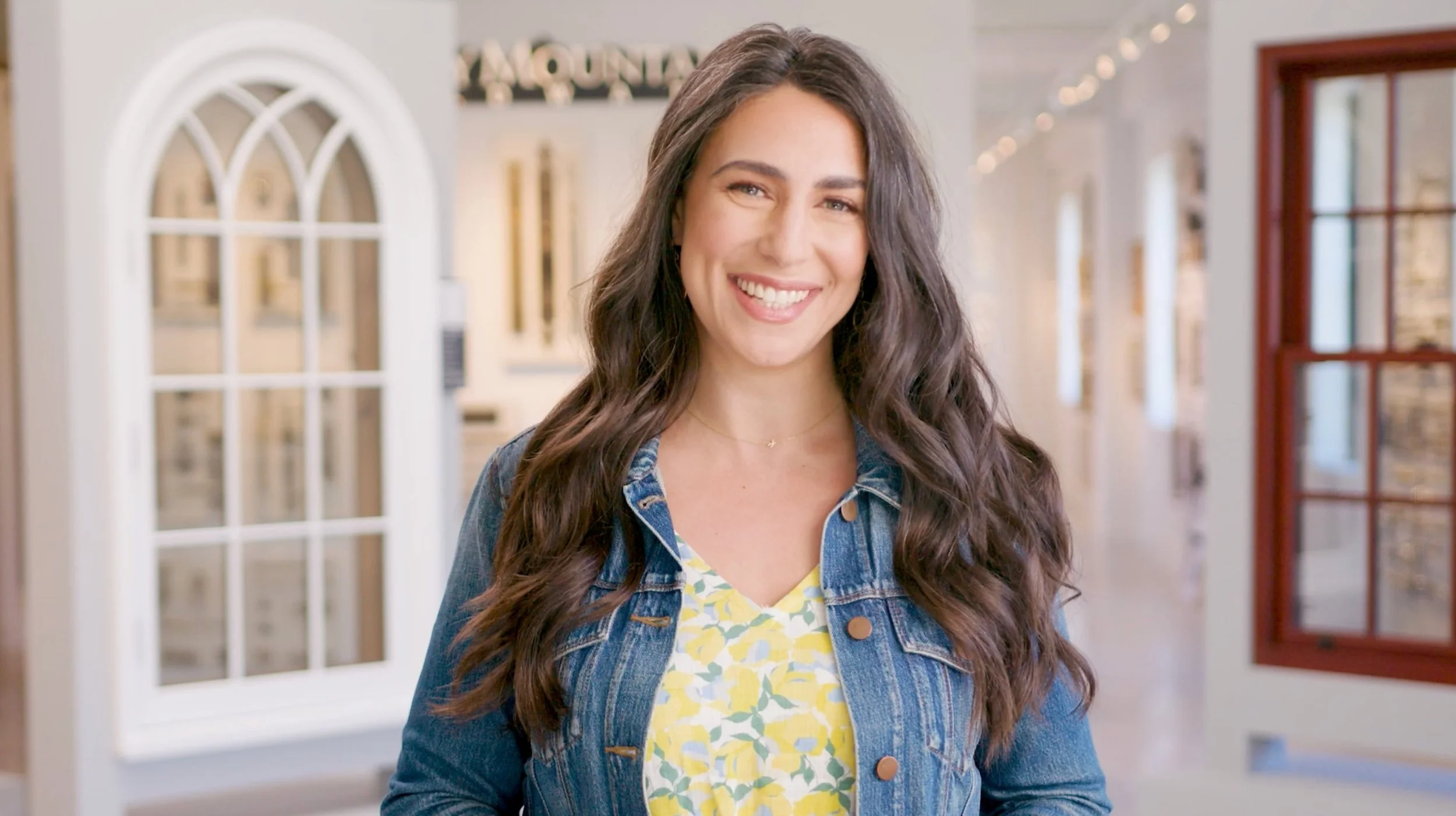 A smiling woman with long wavy brown hair stands indoors, wearing a denim jacket over a yellow floral top, with a background of windows and string lights.