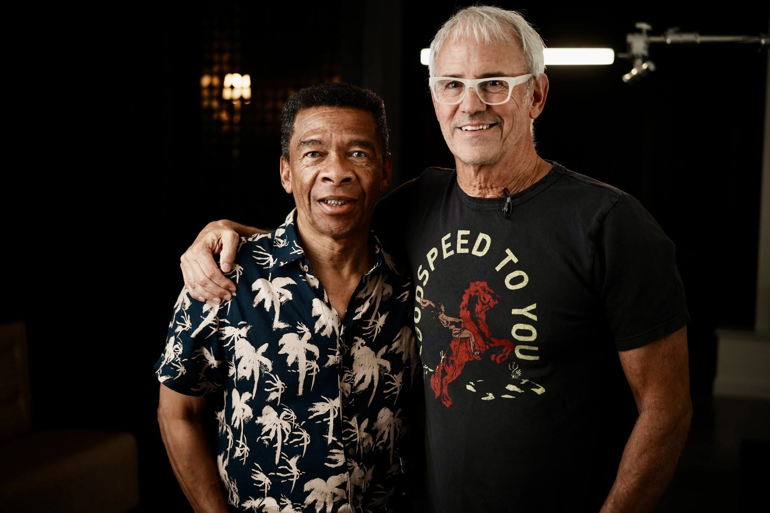 Two men standing closely together, one with his arm around the other, smiling at the camera. The man on the left wears a navy shirt with white palm trees, and the man on the right wears a black T-shirt with a red horse and the text "Good Speed to You