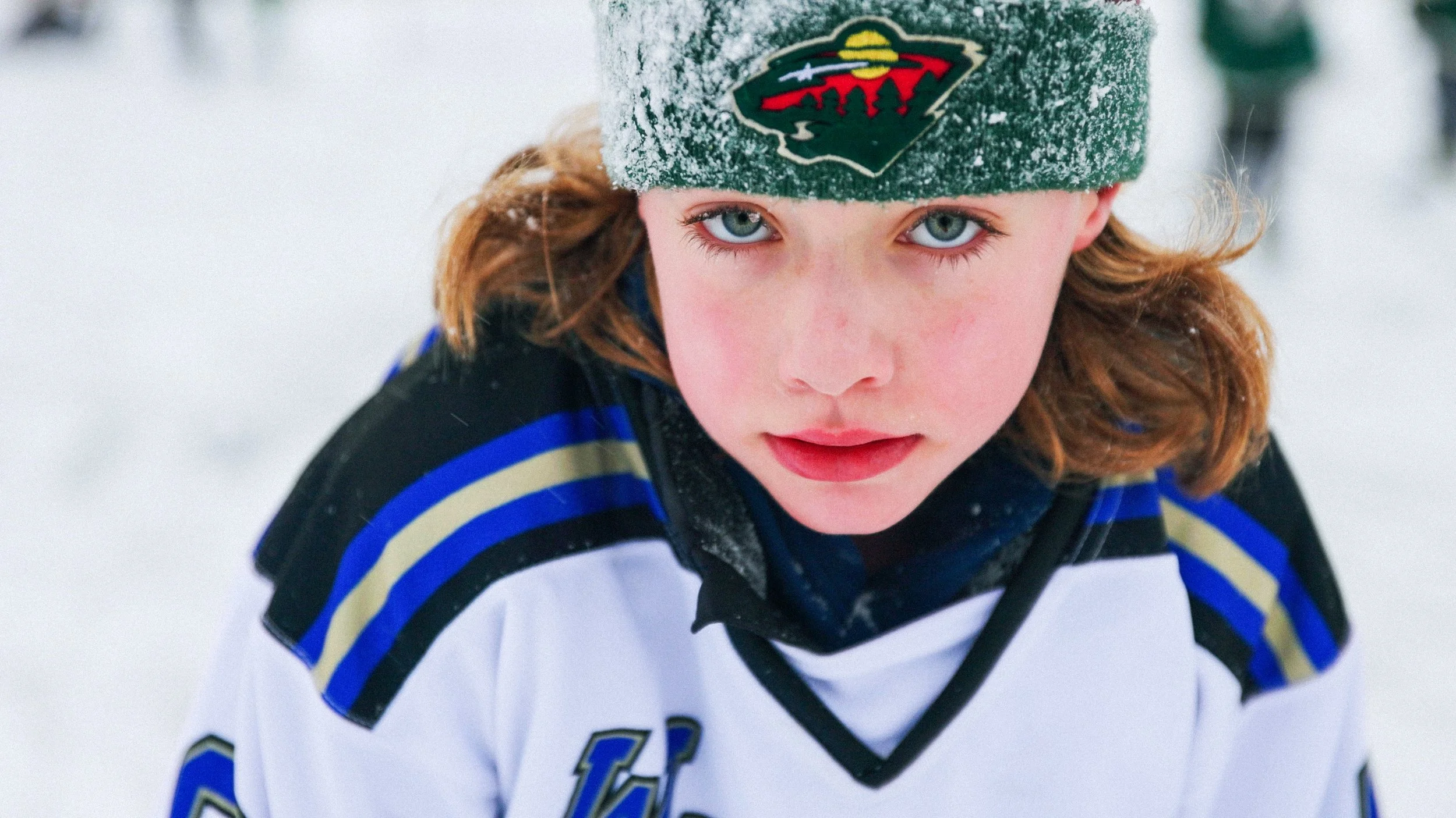 Close-up of a young girl with blue eyes and red hair wearing a Minnesota Wild hockey hat and a hockey jersey, standing outside in the snow.