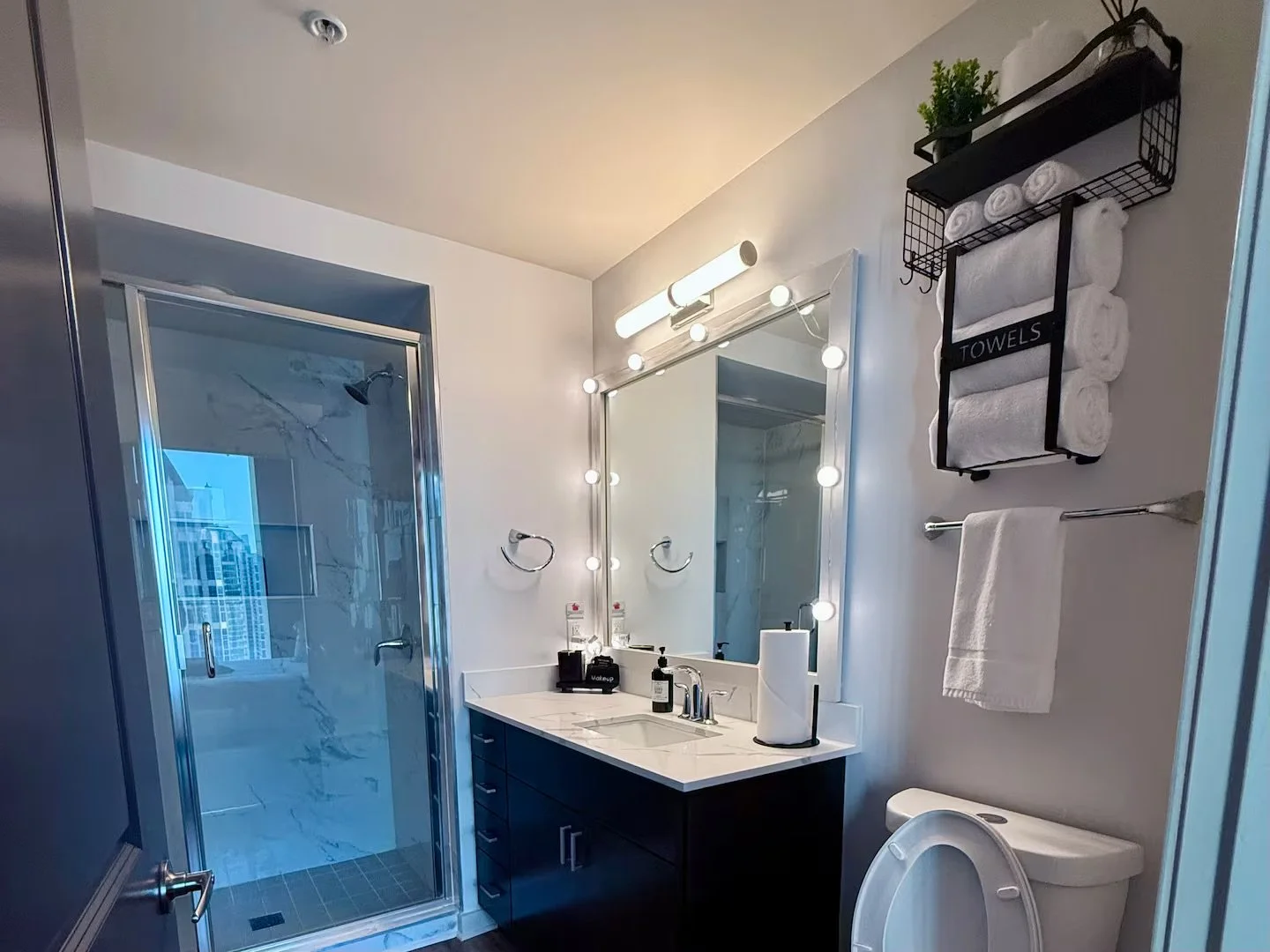 Modern bathroom with a glass shower, black vanity with white countertop, large mirror with lights, wall-mounted towel rack with rolled towels, and a toilet.