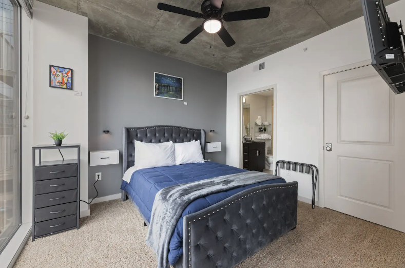 Modern bedroom with gray upholstered bed, white pillows, blue comforter, light gray walls, artwork, small black dresser, black ceiling fan, sliding glass door, and TV mounted on the wall.