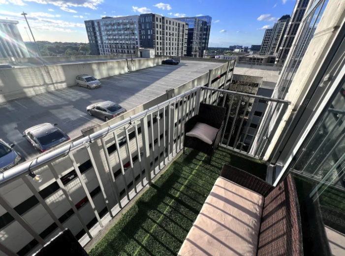 Small balcony with artificial grass, black wicker chair with pink cushion, and a patio chair with striped cushion, overlooking a parking garage and modern buildings against a blue sky.